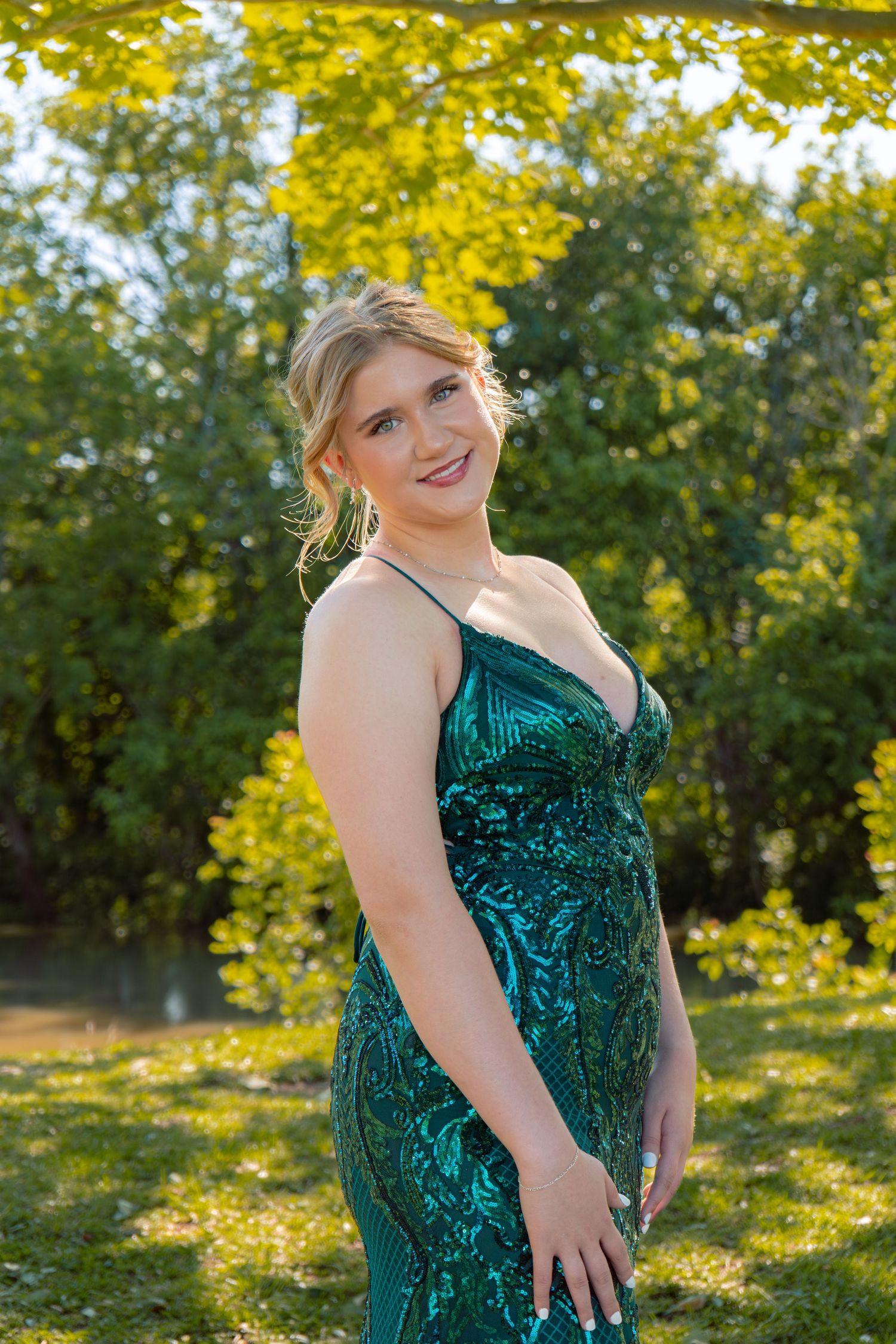 Smiling blonde teen in emerald sequin prom dress poses in sunny park.
