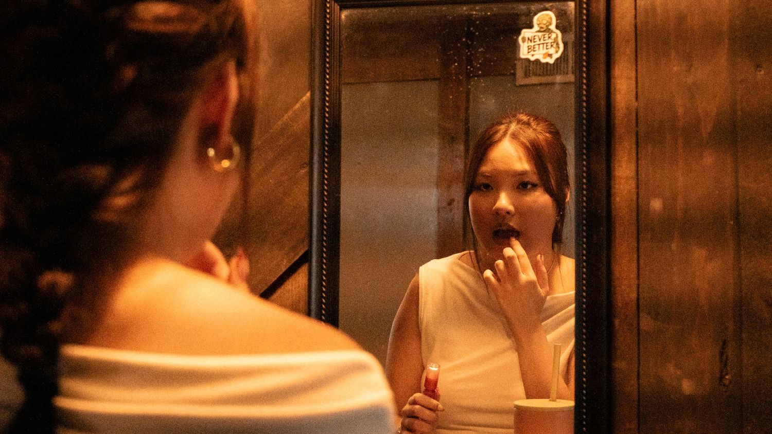 Young woman examining her teeth in an ornate gold-framed mirror in a dimly lit room.