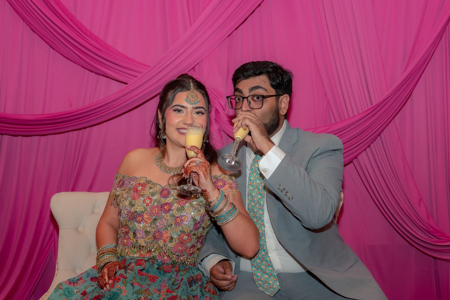 Couple in festive attire drinking champagne in front of pink draped backdrop at Indian celebration.
