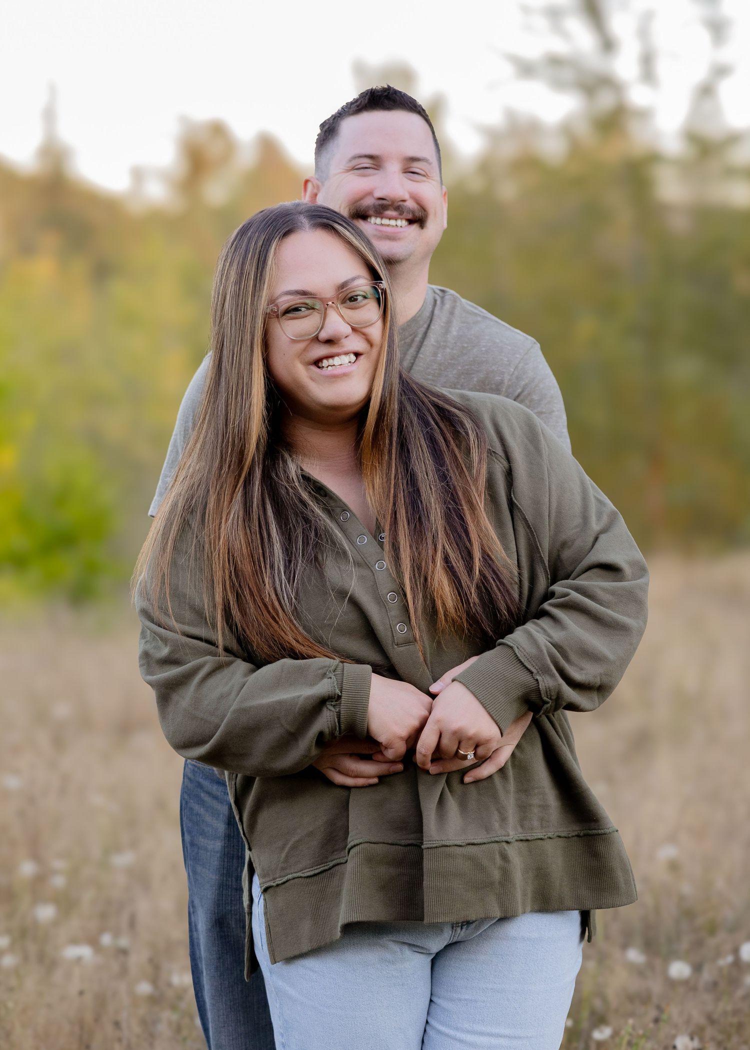 Joyful couple embracing in an open field, woman in green hoodie smiling brightly.