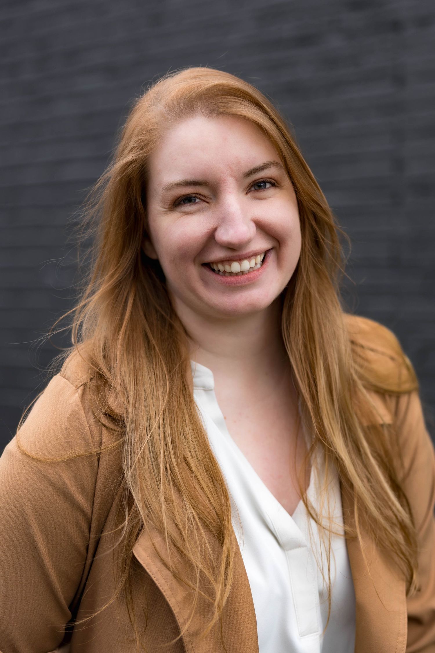 Happy redhead woman in tan blazer and white blouse smiling against a dark wall.