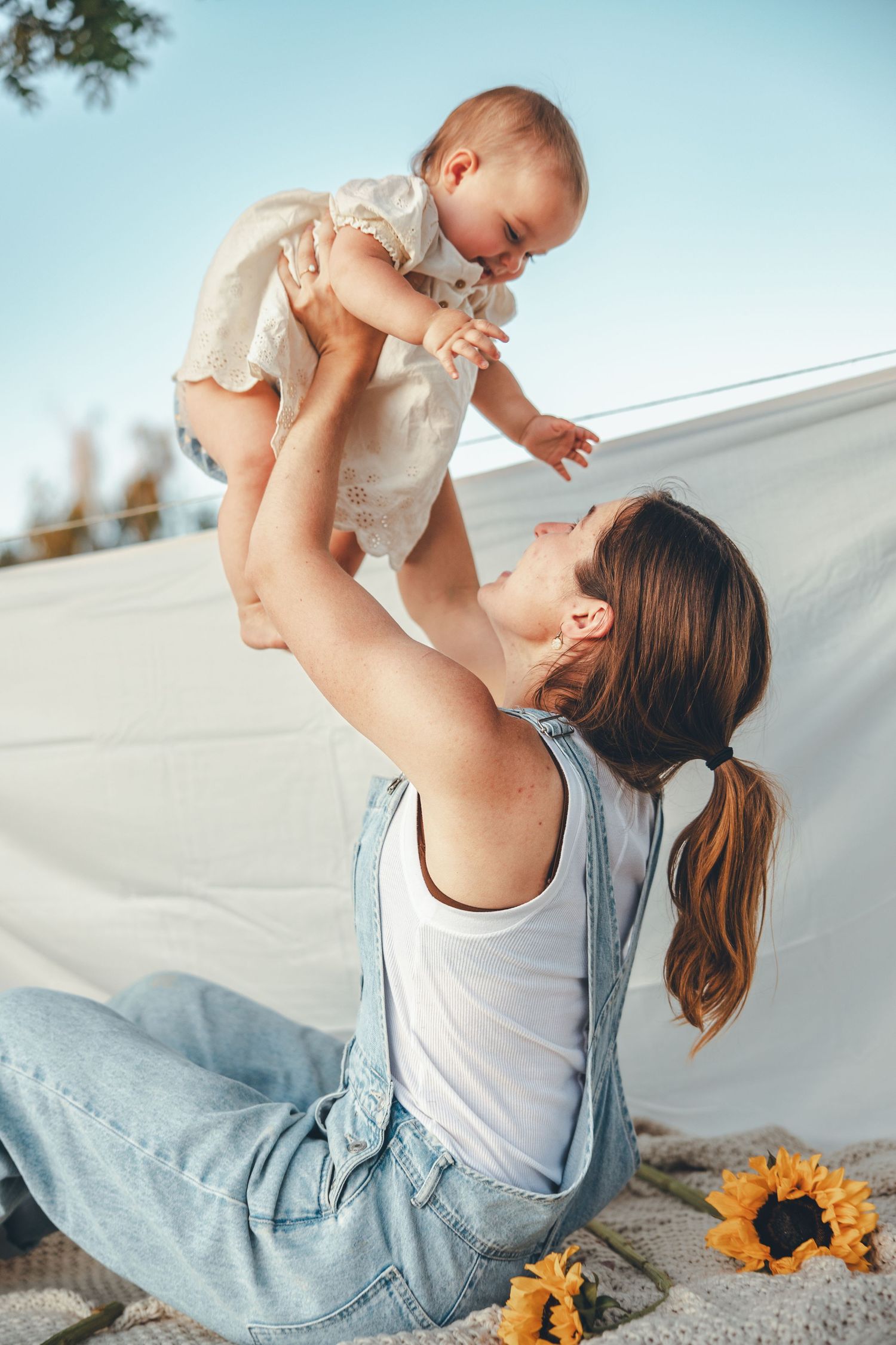 Mother sitting outdoors lifting smiling baby in white dress against blue sky with sunflowers nearby.