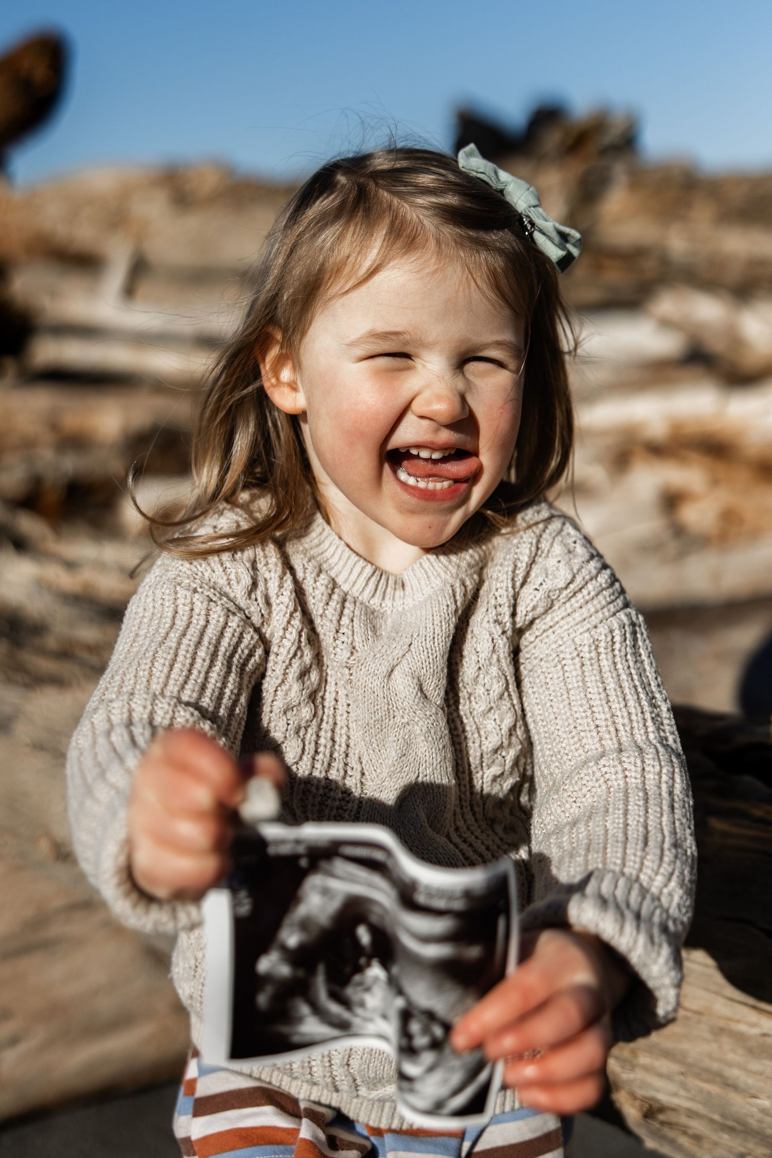 Excited toddler girl in knit sweater holding ultrasound photo outdoors, announcing a new sibling.
