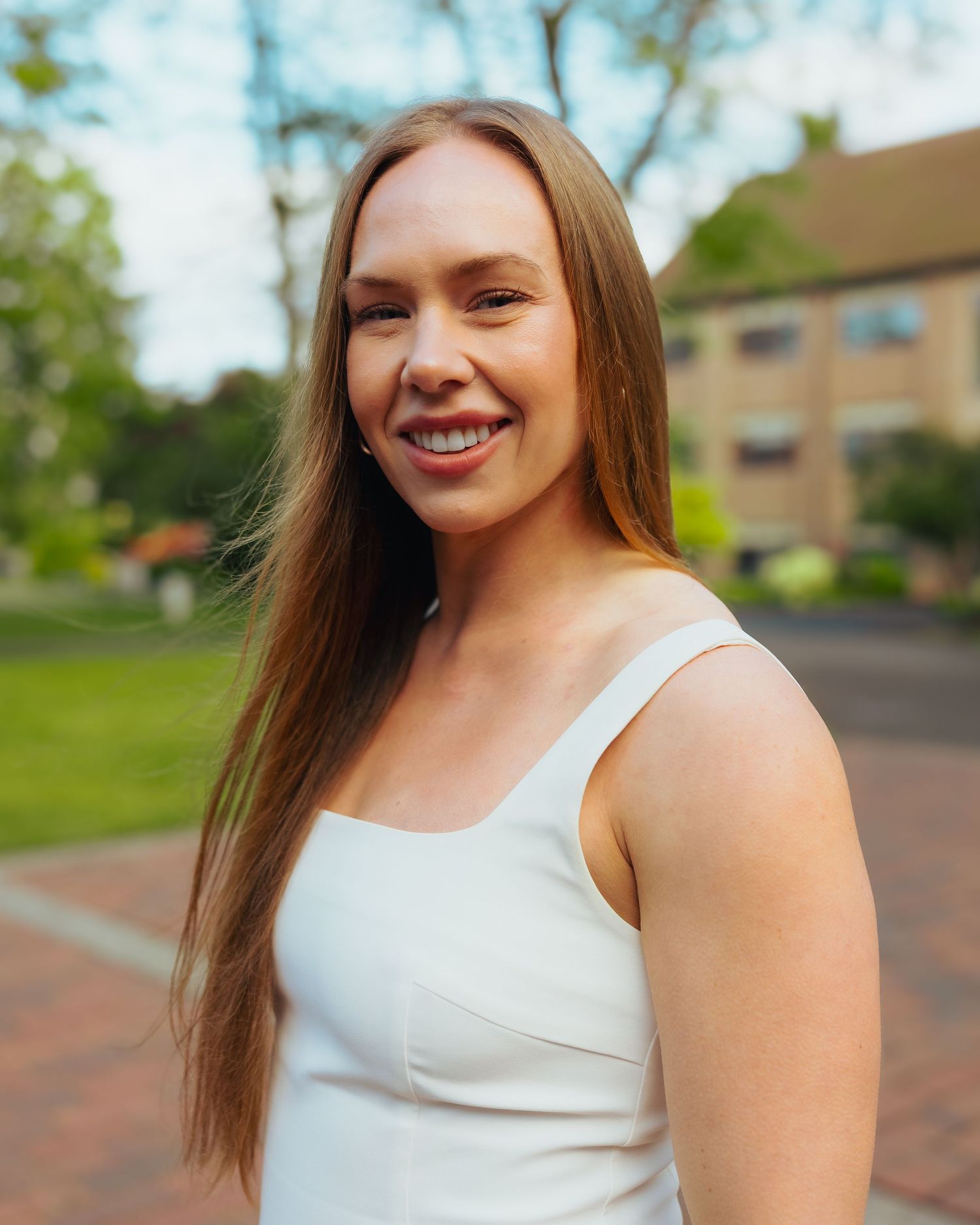Smiling young woman with long auburn hair wearing a white tank top outdoors near a brick pathway.