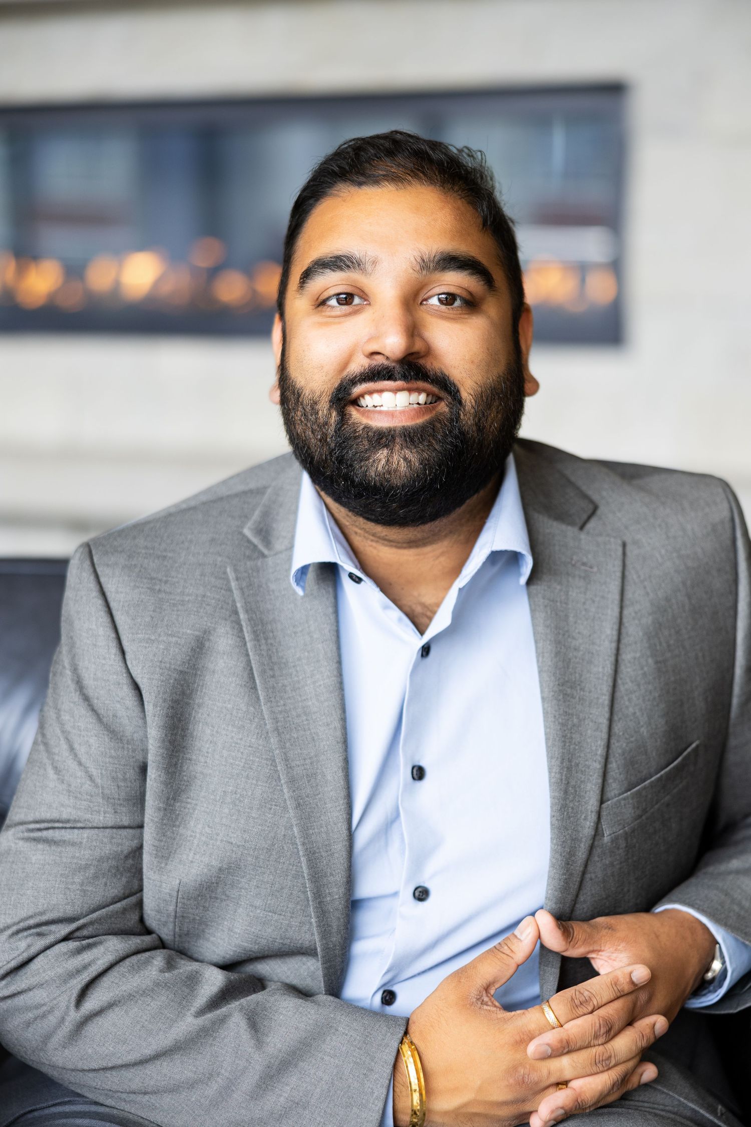 Smiling bearded man in gray blazer and blue shirt seated near modern fireplace in professional setting.