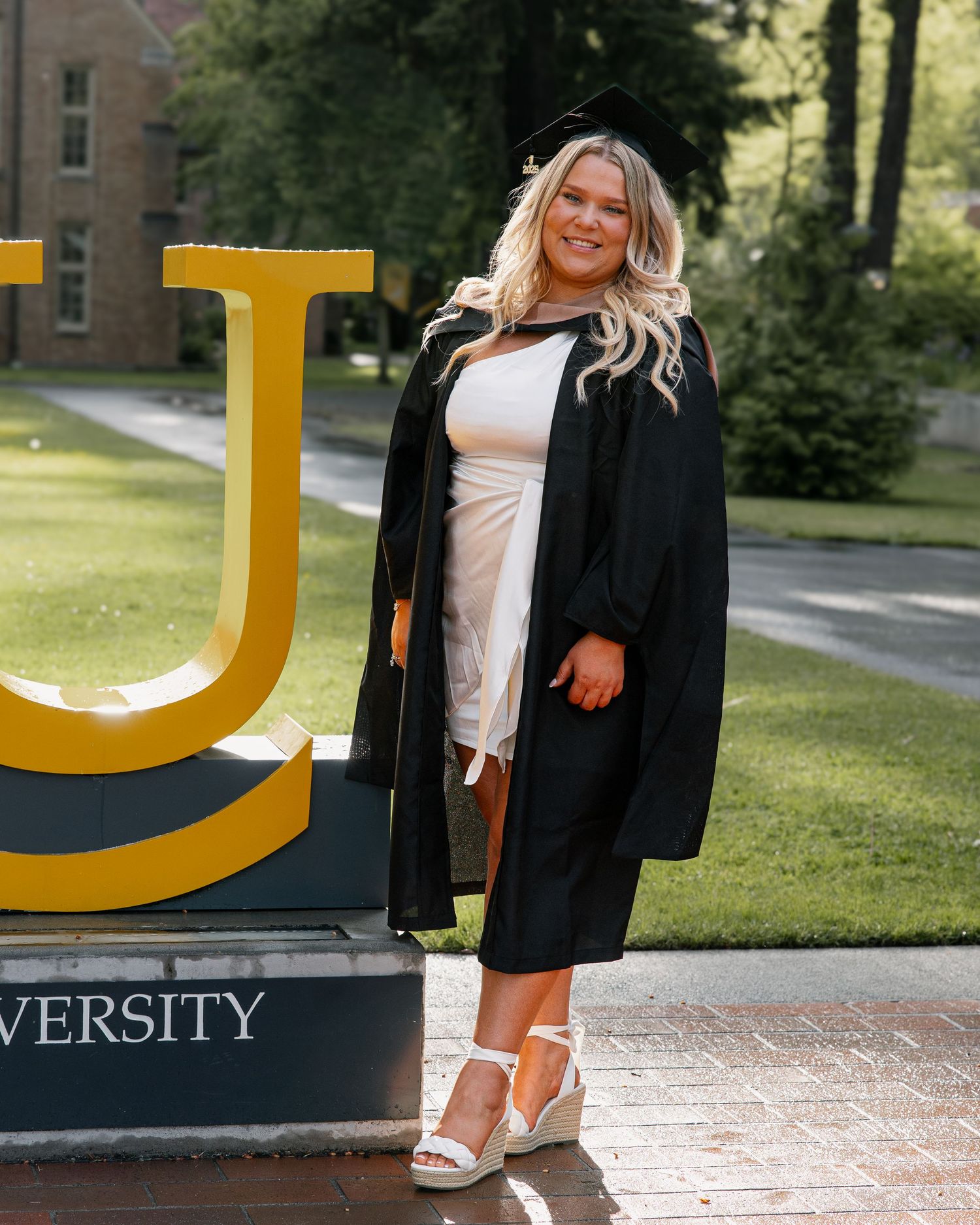 Female graduate in black cap and gown poses in front of a university letter sign on a green campus.