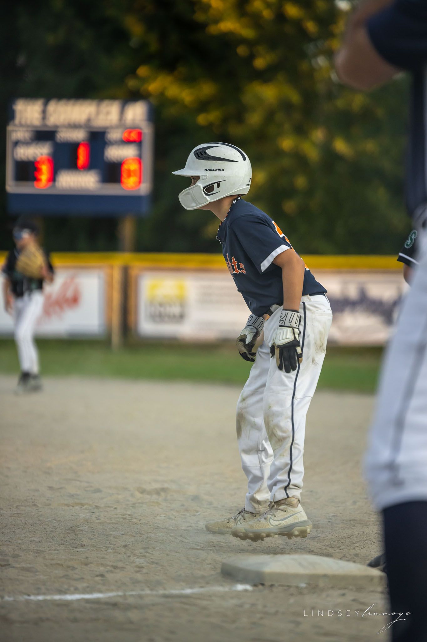 Youth baseball player in white uniform and helmet stands on base during an evening little league game.