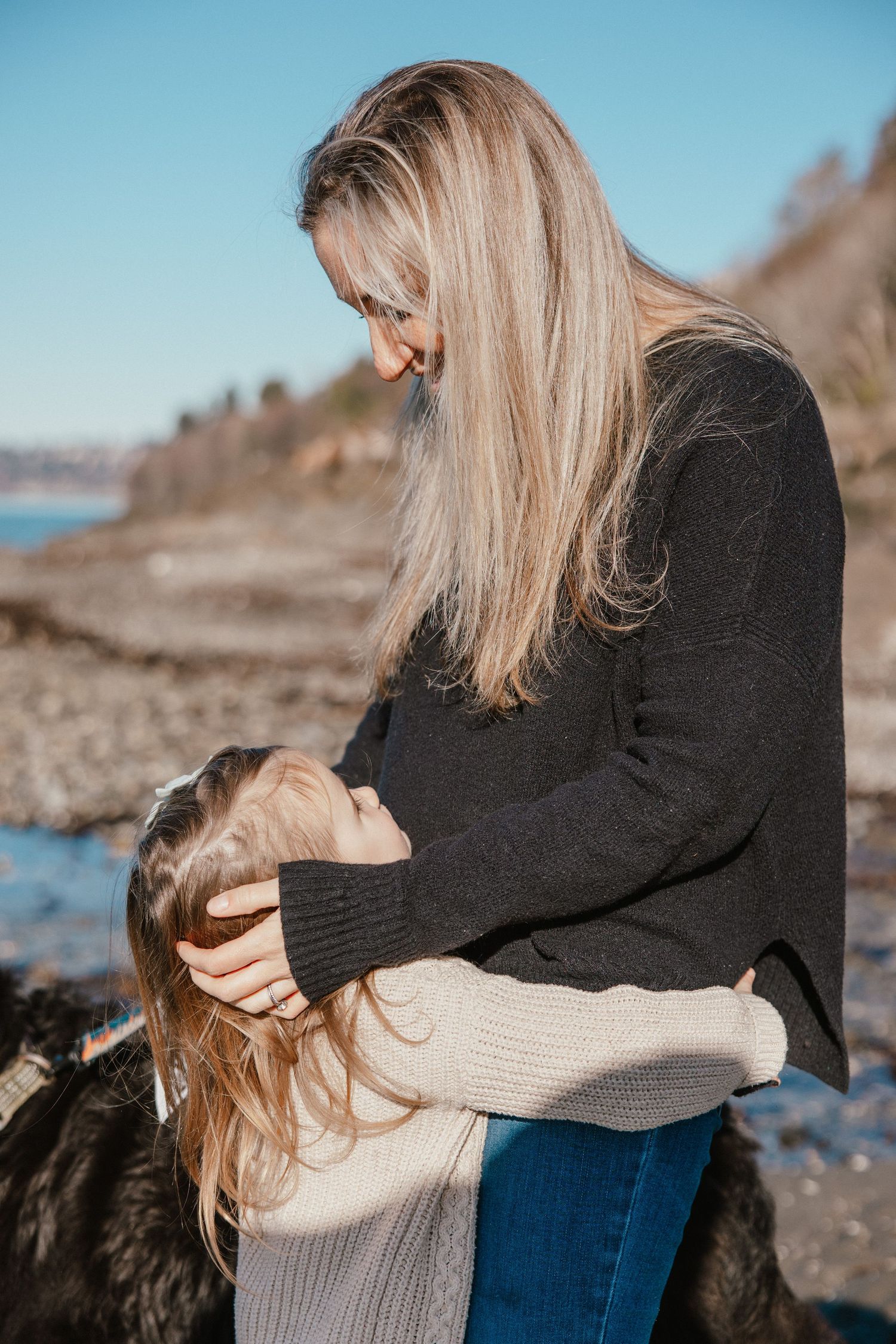 A mother tenderly holds her young daughter on a rocky beach, both wearing cozy sweaters on a sunny day.