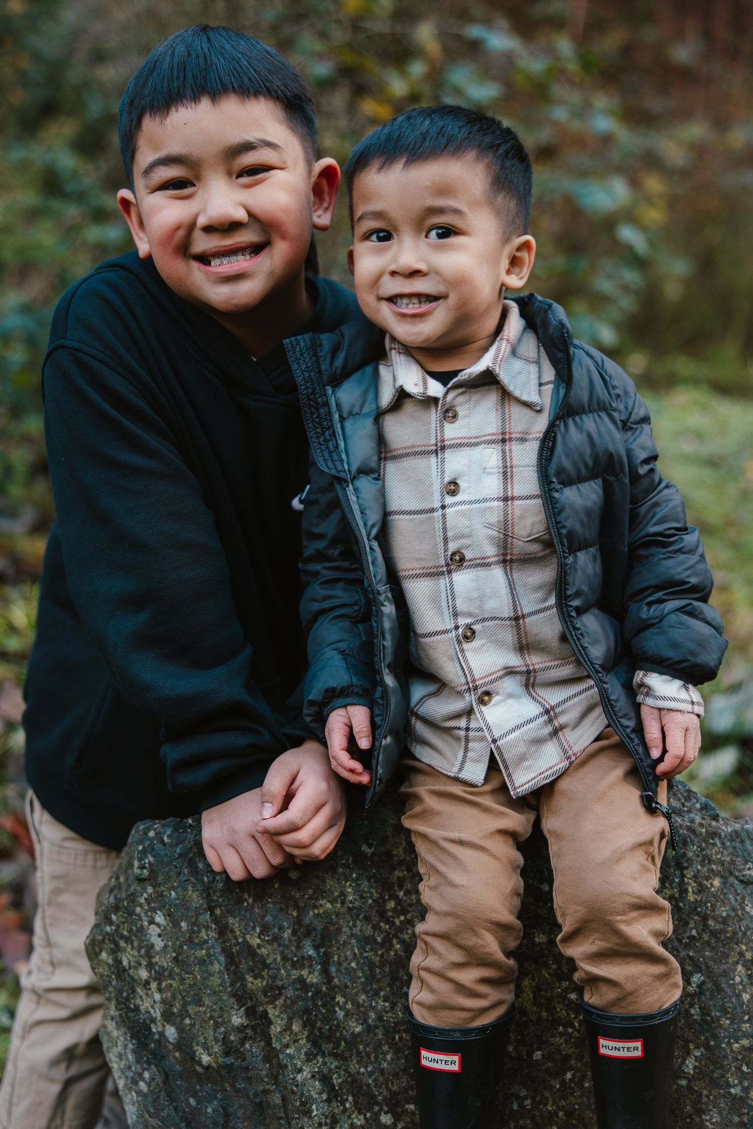 Two young boys smiling outdoors on a rock, wearing casual jackets and khaki pants in a forest setting.