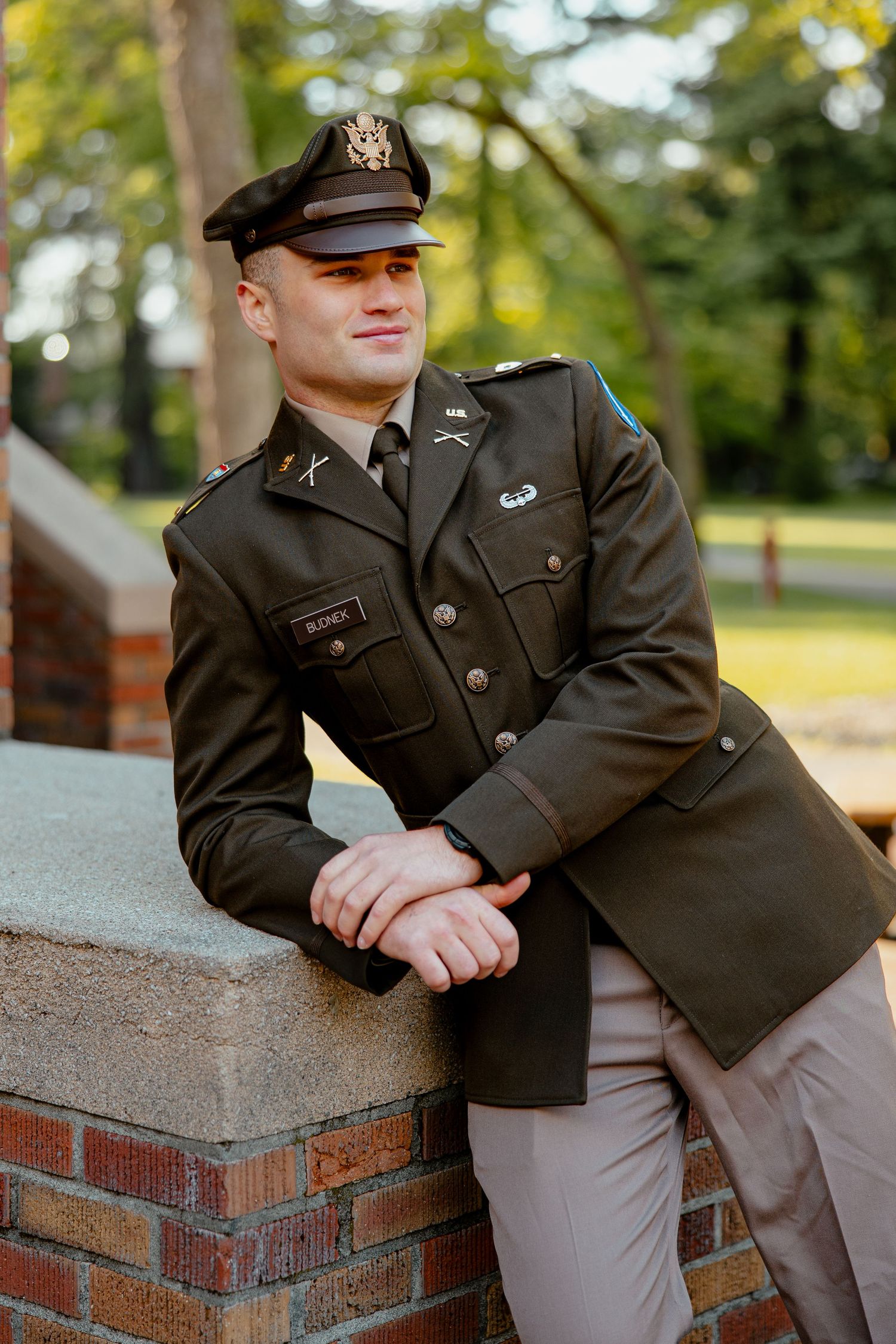 Young man in US Army dress uniform leaning on brick wall outdoors, smiling confidently.