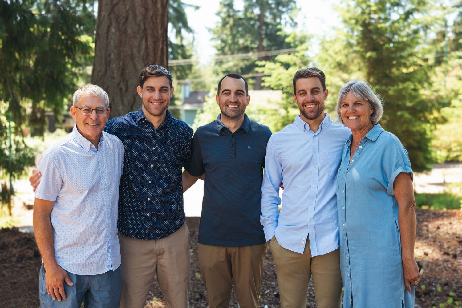 Family of five smiling outdoors among tall trees on a sunny day.