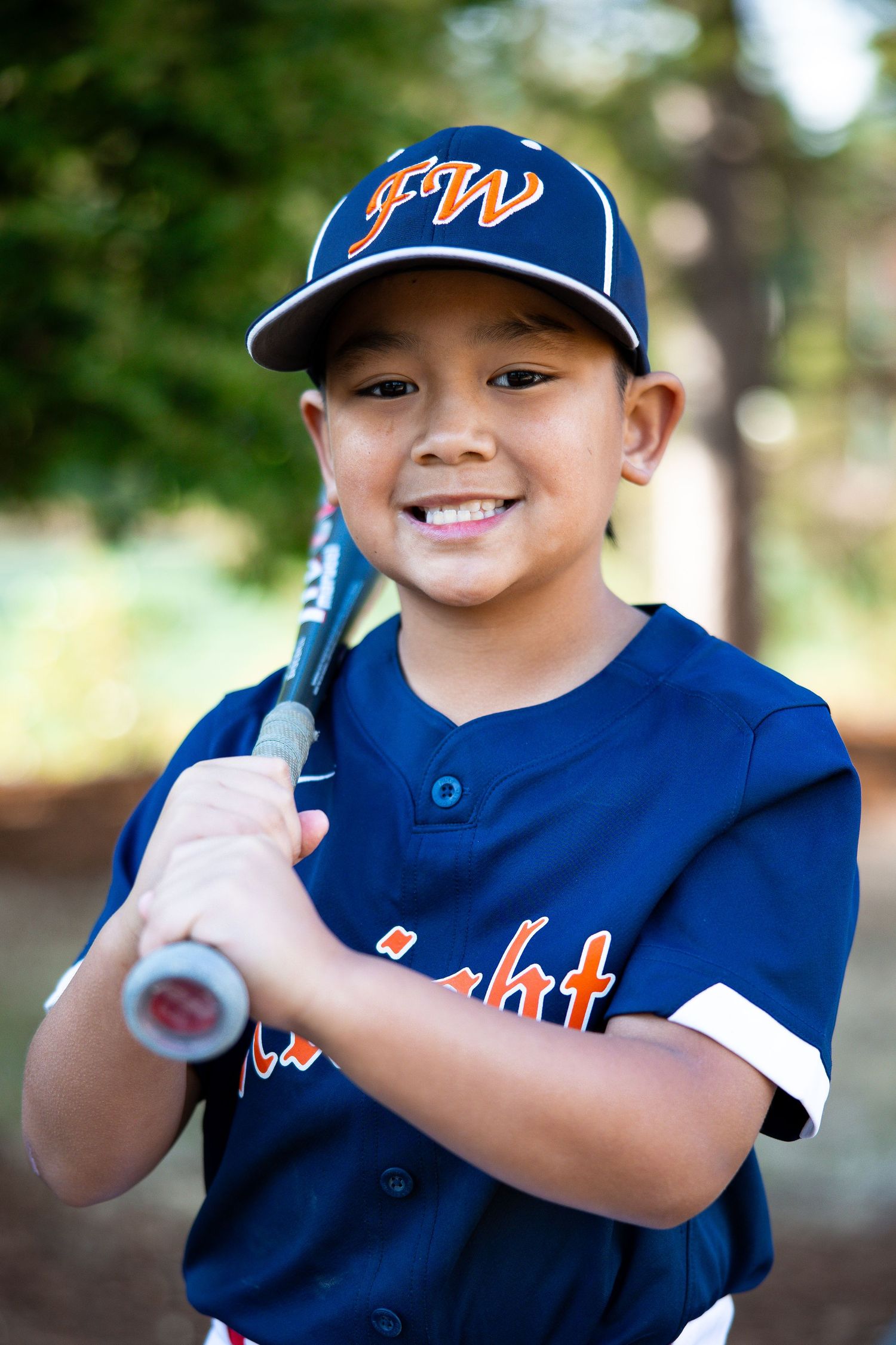 Smiling young boy in navy FW Knights baseball jersey holds a bat for an outdoor portrait.
