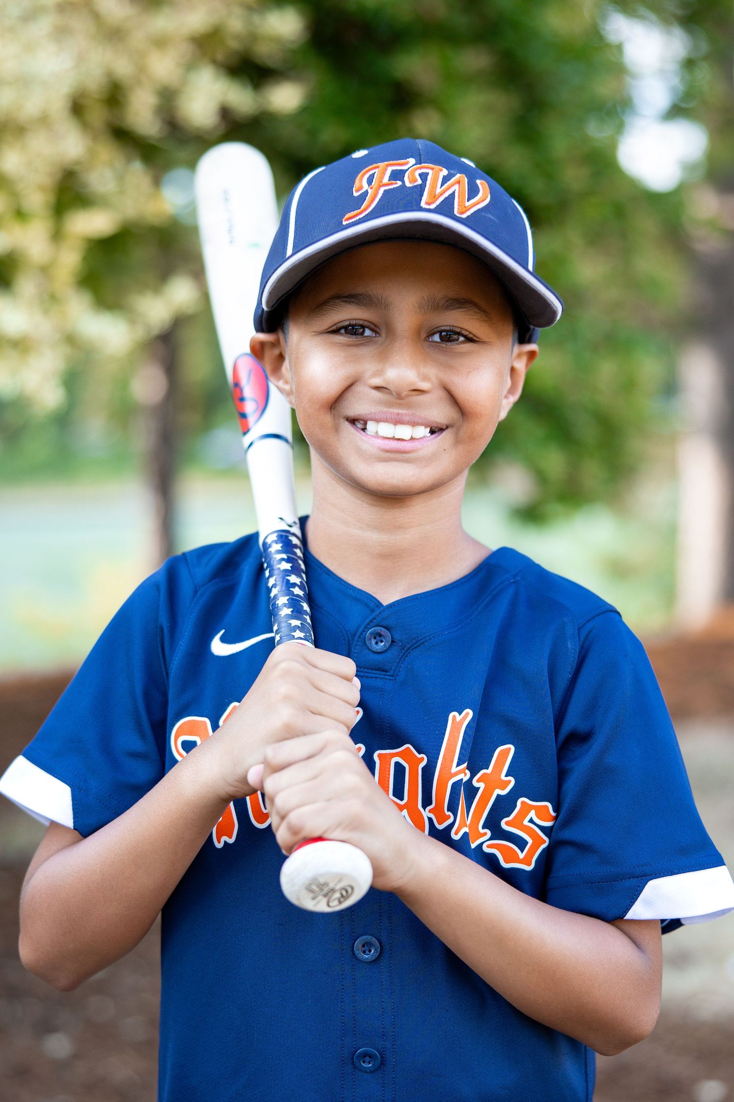 Smiling young boy in navy FW Knights baseball jersey holds a bat for an outdoor portrait.