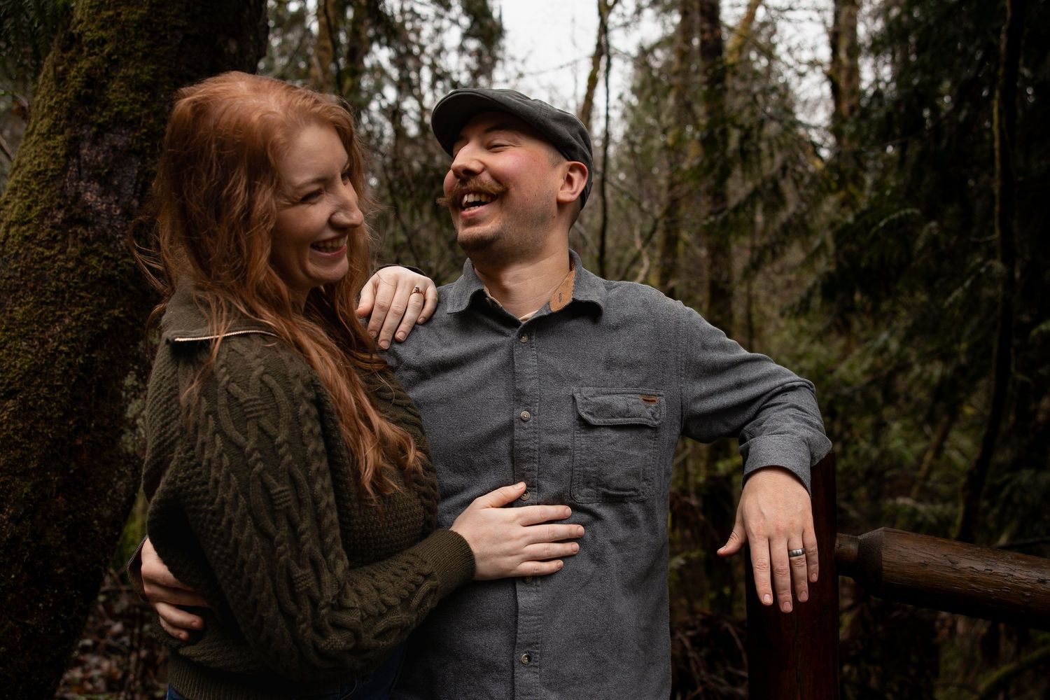 Laughing redhead woman embraces smiling man in gray shirt among mossy forest trees.
