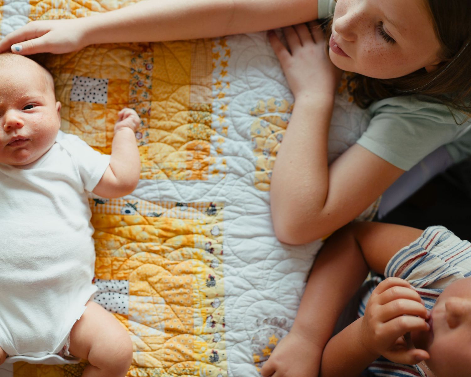 Young girl lying on yellow quilt tenderly touching newborn baby's head from above.