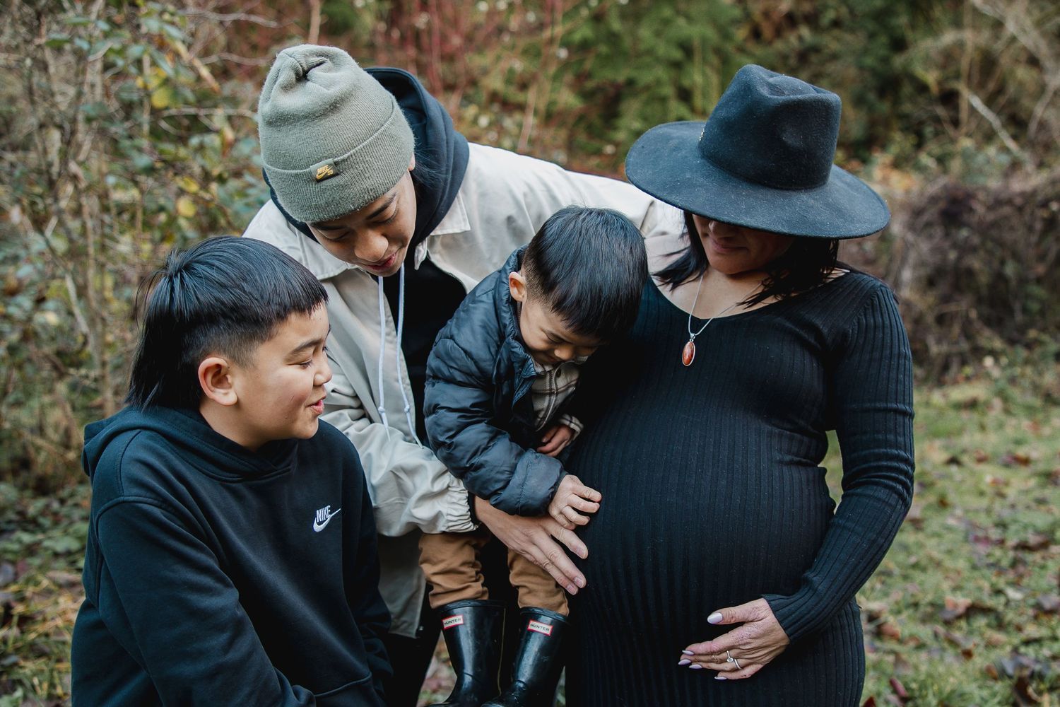 Pregnant woman in black dress with family outdoors, man in Nike hoodie and child in winter setting.