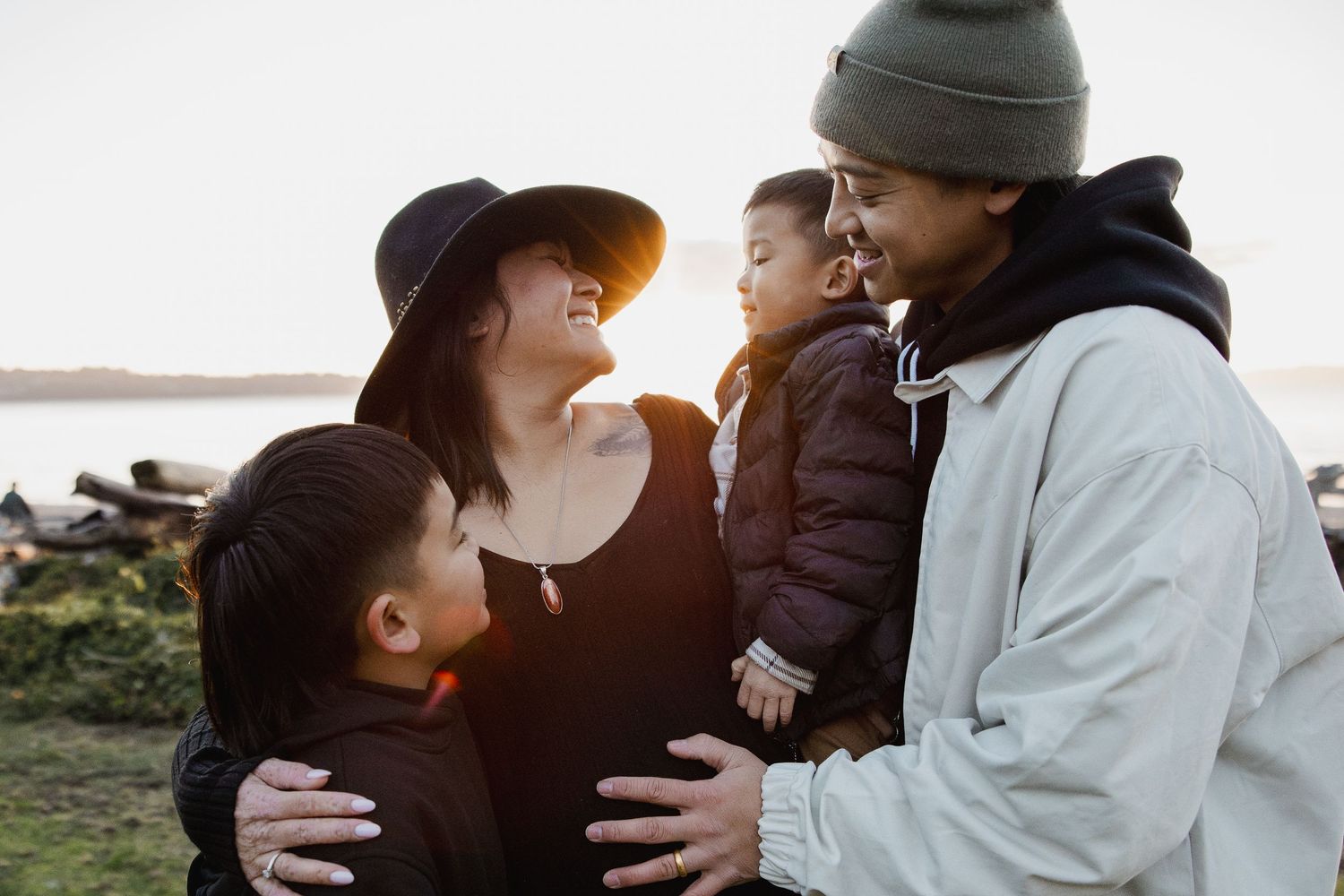 Happy family of four enjoying a sunset outdoors near water, parents holding young children while smiling.