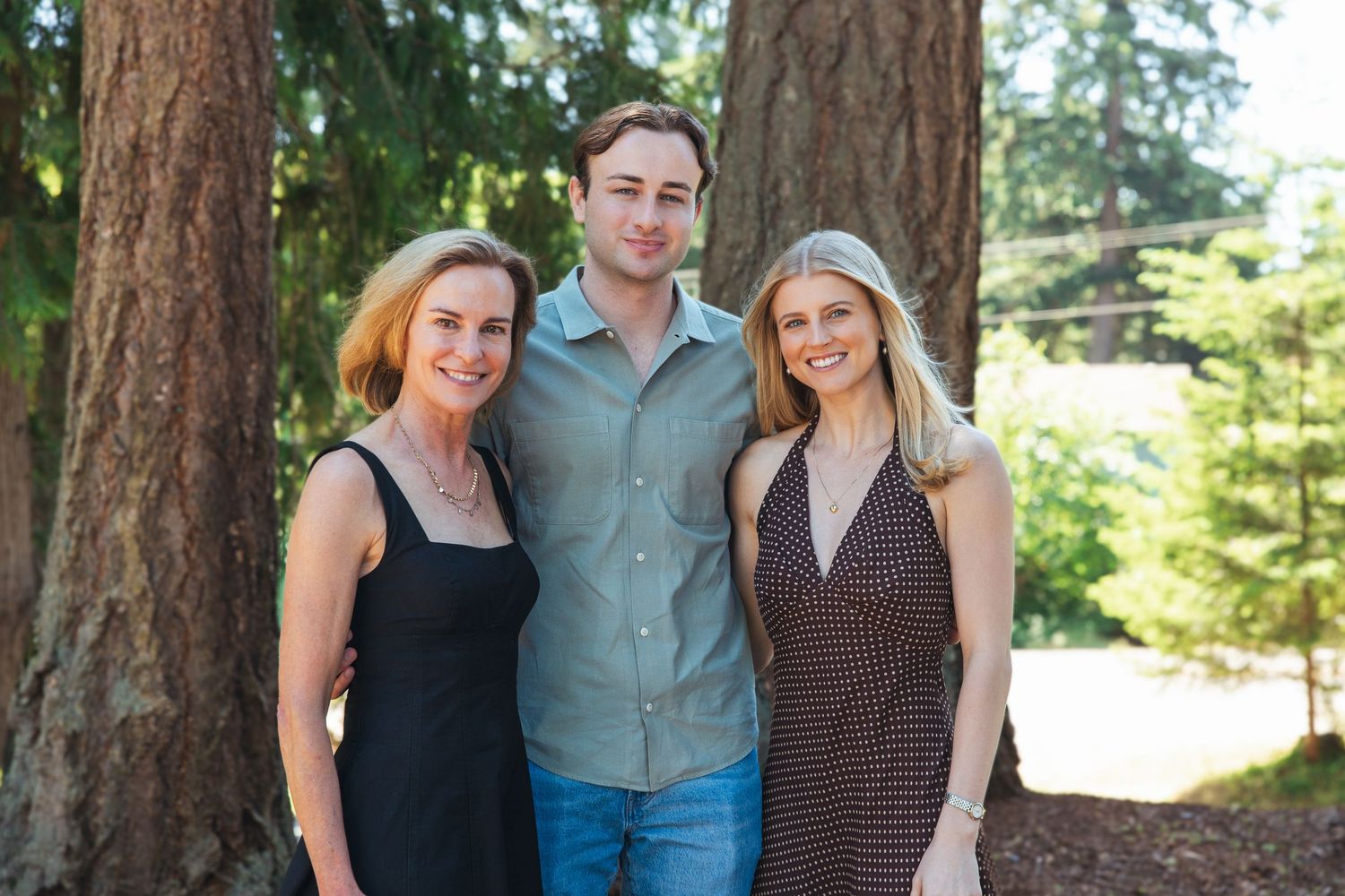 Two women and a man smiling together outdoors near tall trees.