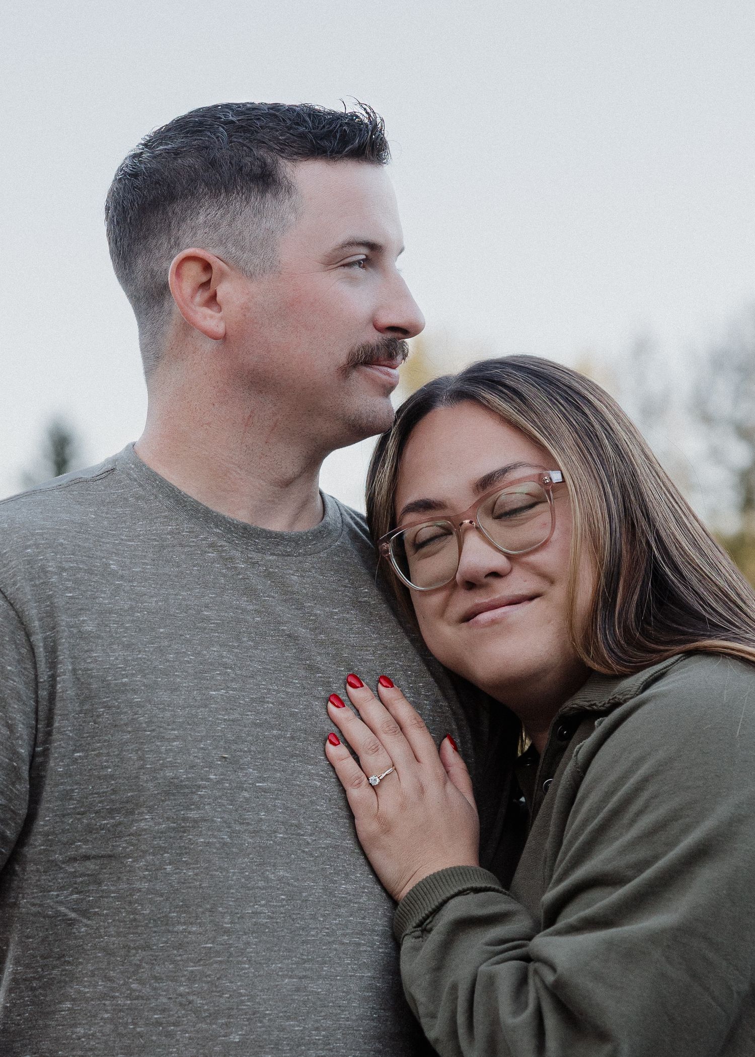 Couple cuddling outdoors at dusk, woman with glasses and engagement ring leaning against mans chest contentedly.