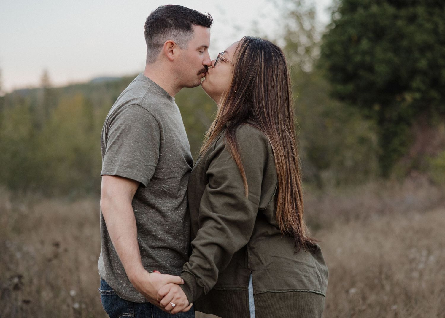 Couple sharing a kiss while holding hands in a natural field setting during engagement photos.