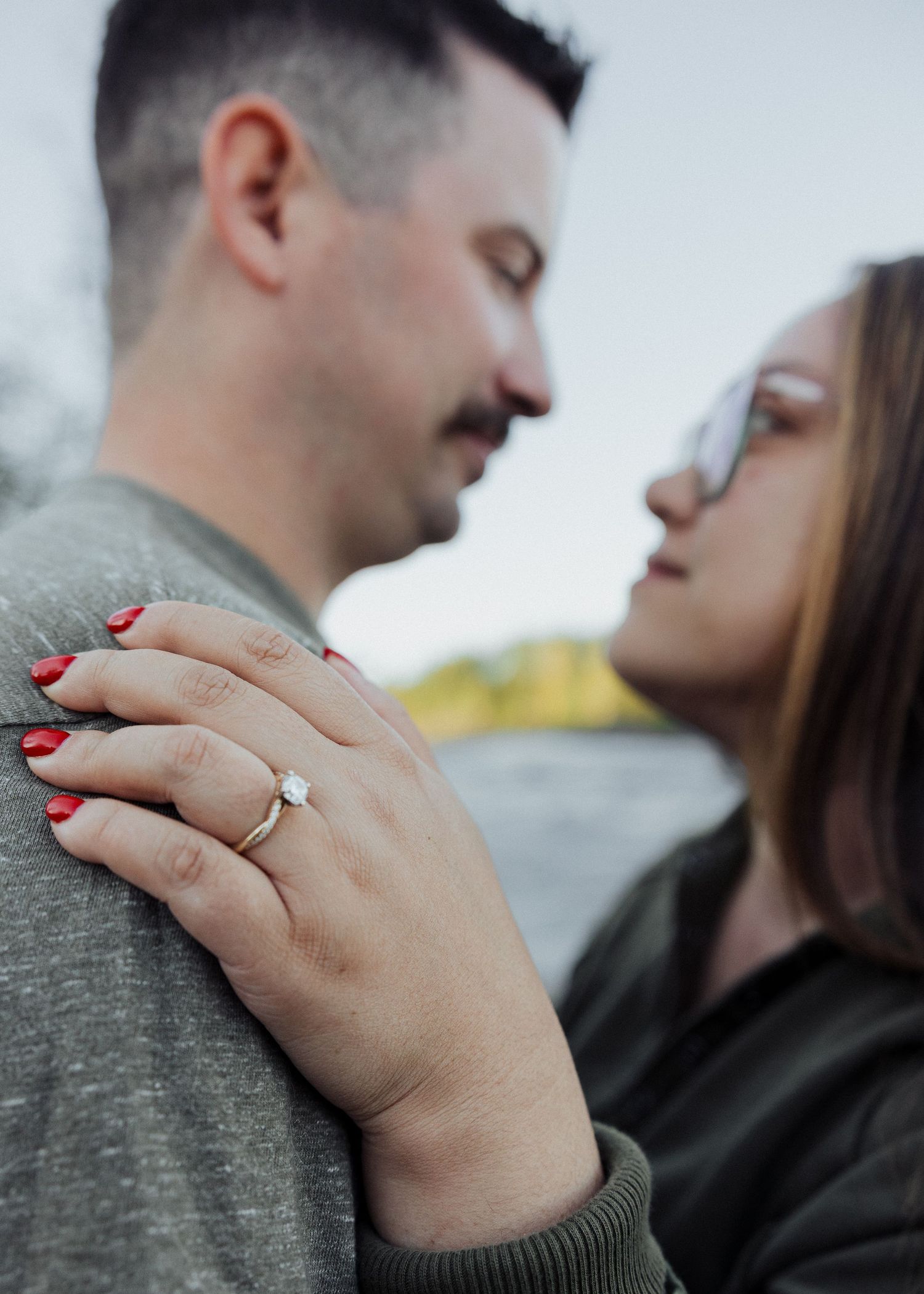 Close-up of woman with red nails showing engagement ring while couple shares intimate moment outdoors.
