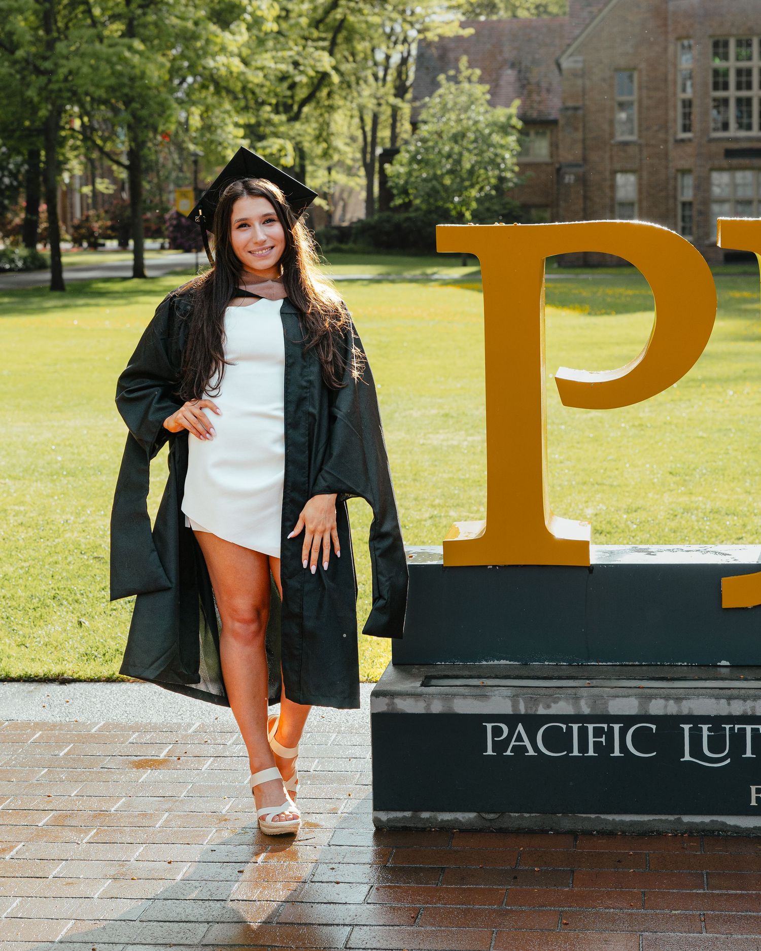 Graduate in cap and gown posing by Pacific Lutheran University sign outdoors.