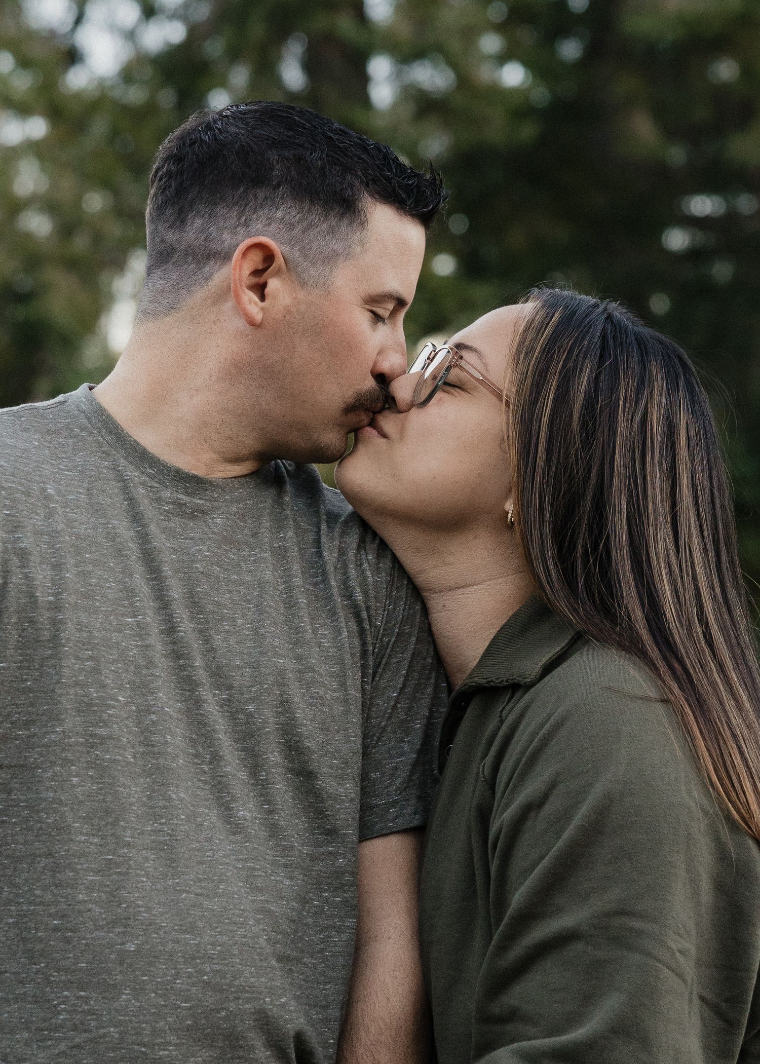 Engaged couple sharing a romantic kiss outdoors with green trees in the background.