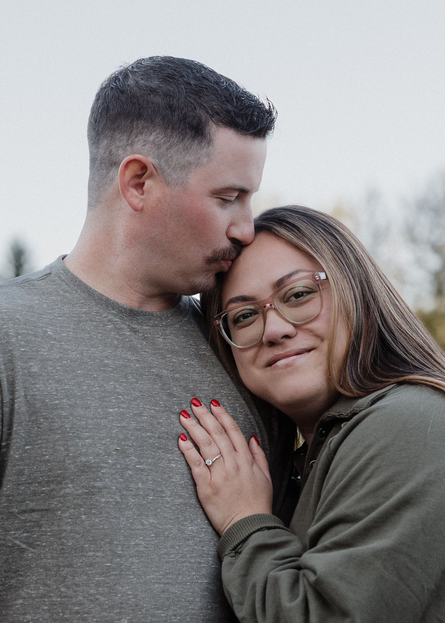 Man kissing womans temple as she smiles, woman wearing glasses and engagement ring with red nails.