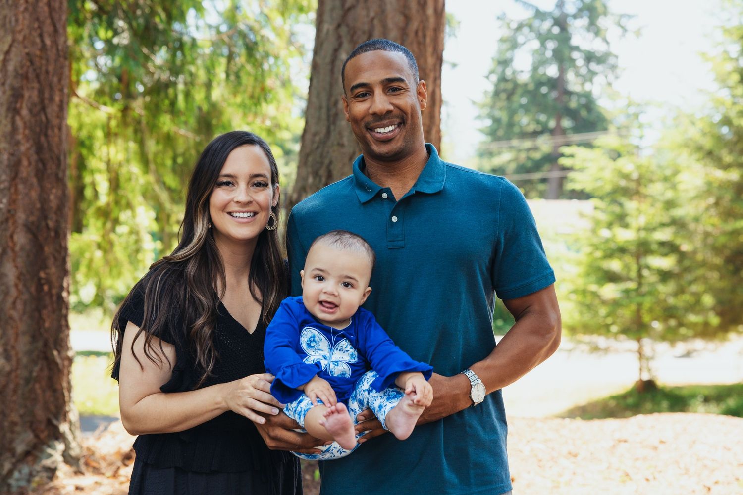 Smiling couple holding their happy baby boy outdoors near tall trees.