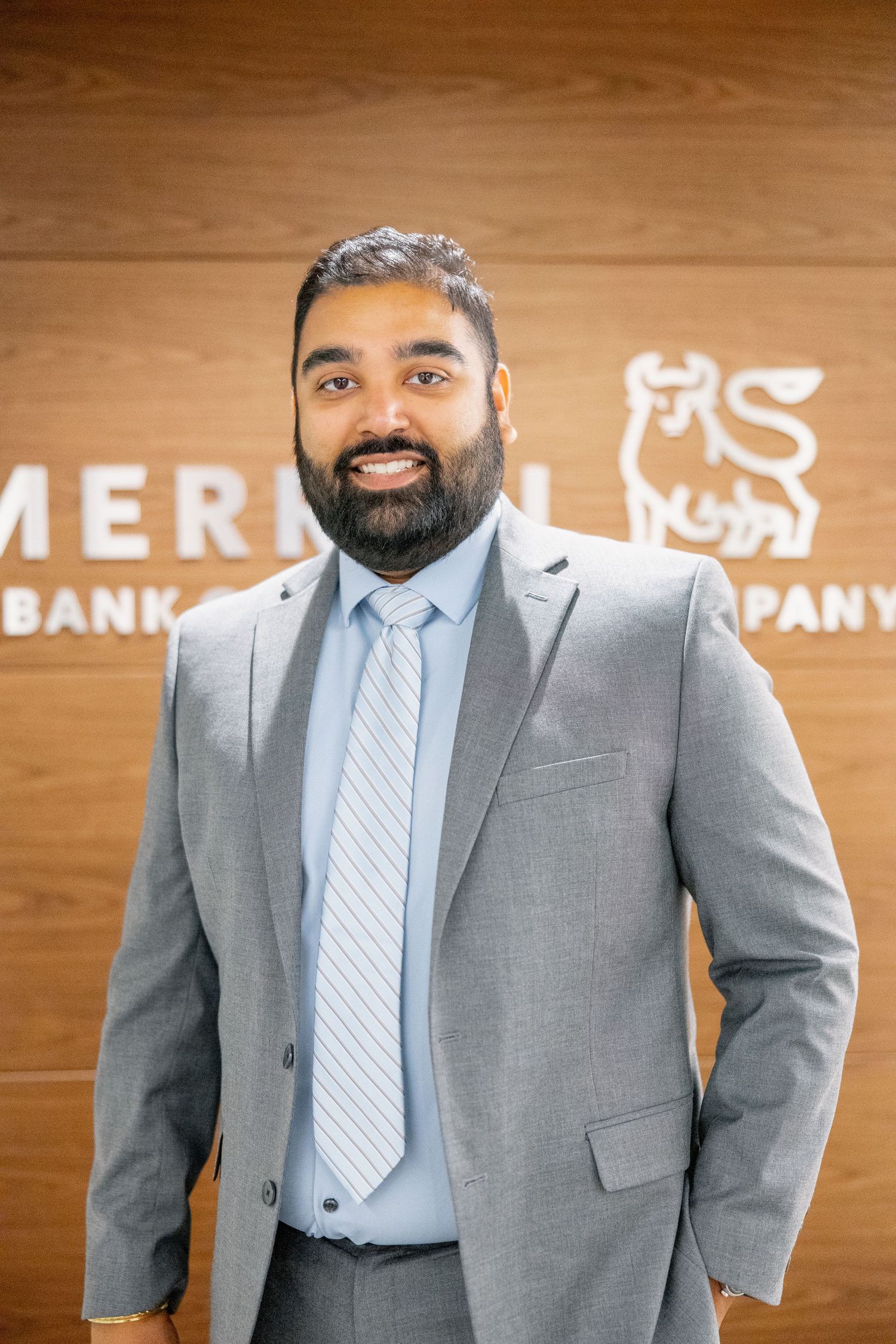 Bearded South Asian man in gray suit smiling confidently in front of Merrill Lynch wooden logo wall.