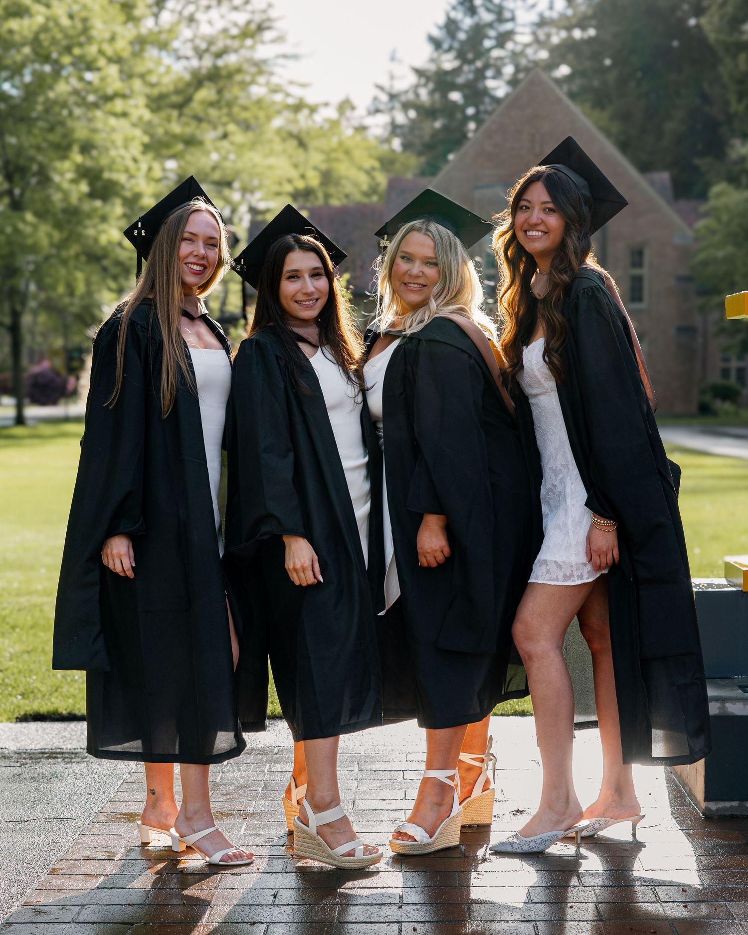 Four female graduates wearing black caps and gowns smile together on a sunny college campus brick pathway.