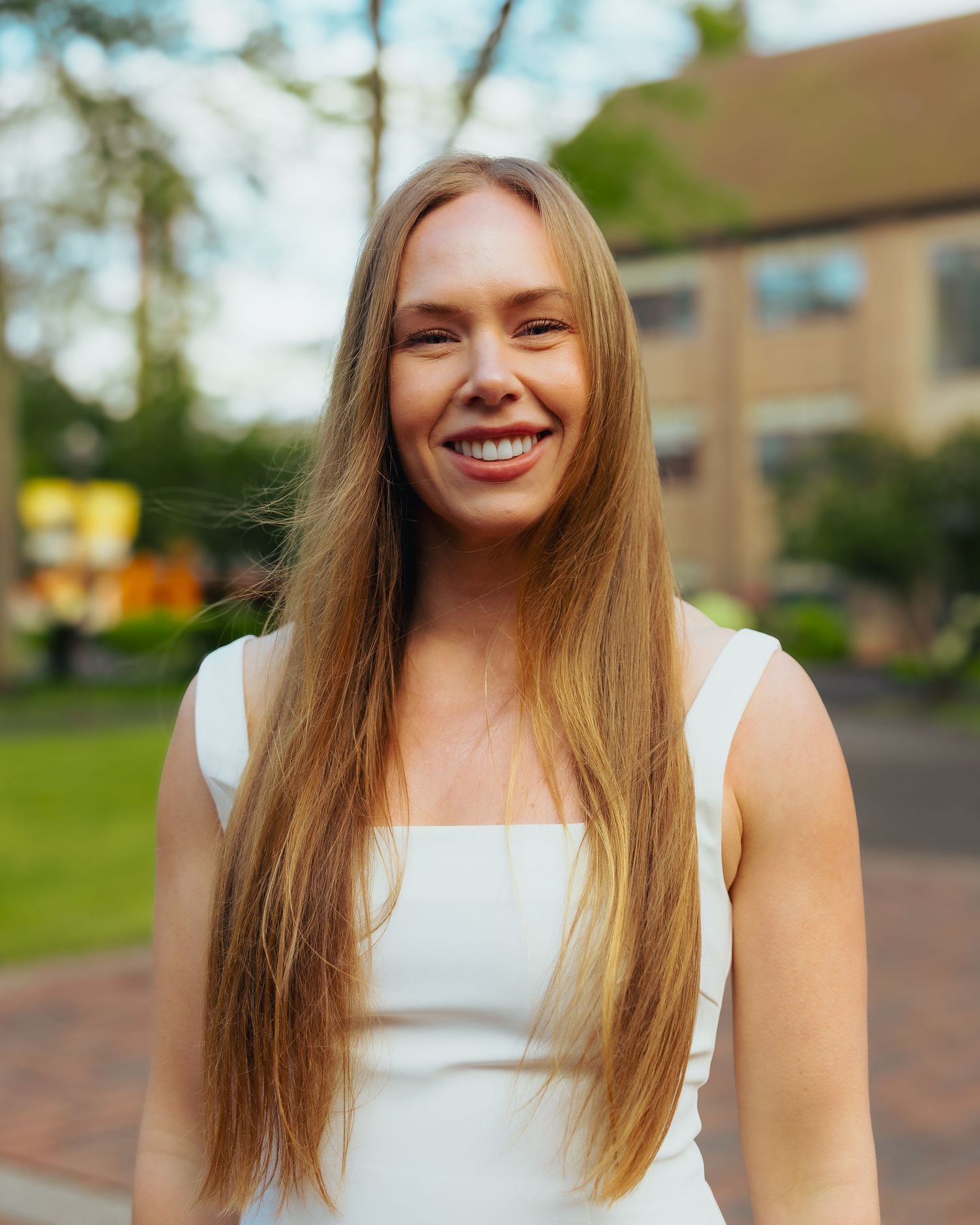 Young woman with long straight blonde hair smiling in a white dress outdoors.