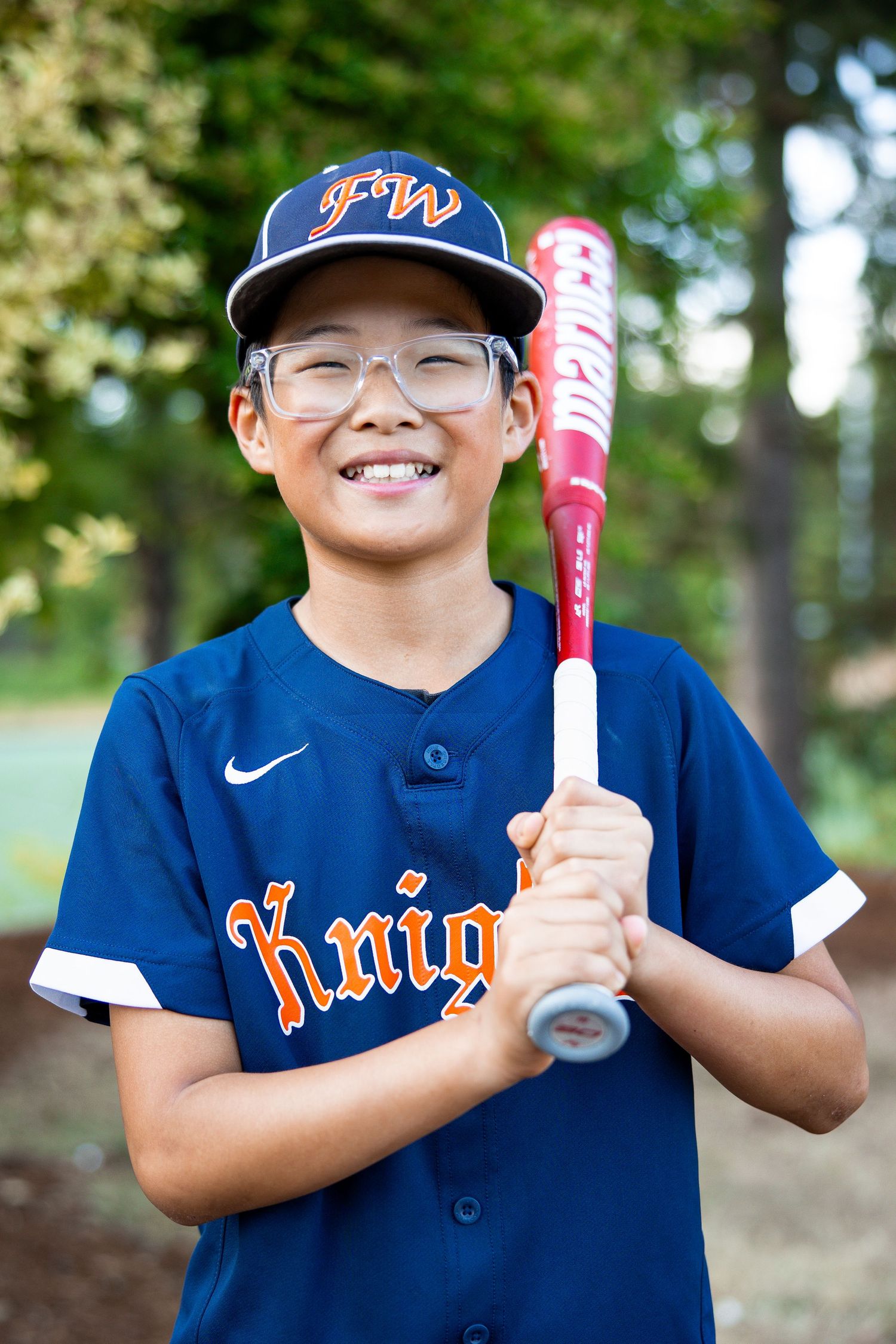 Smiling boy wearing glasses in navy FW Knights uniform holds a red and white baseball bat outdoors.