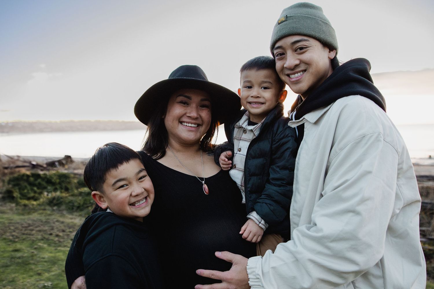 Pregnant woman in black dress and hat smiling with family near waterfront at sunset.