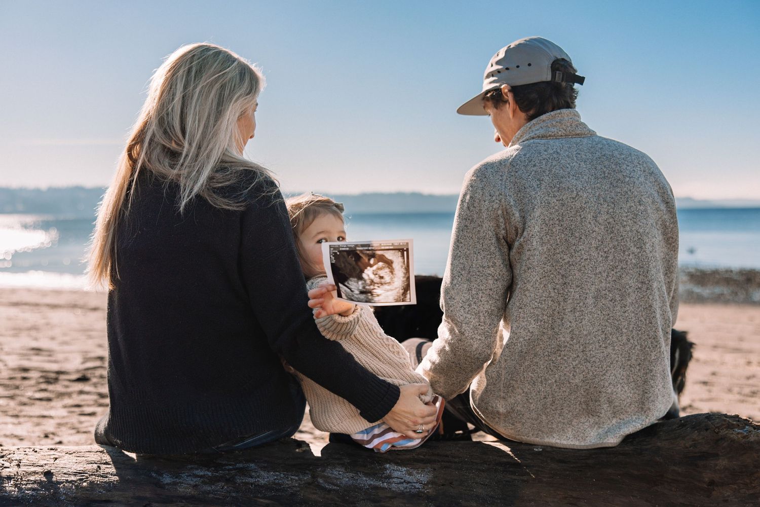 Family of three sitting on a beach rock viewing a printed photo together on a sunny day.