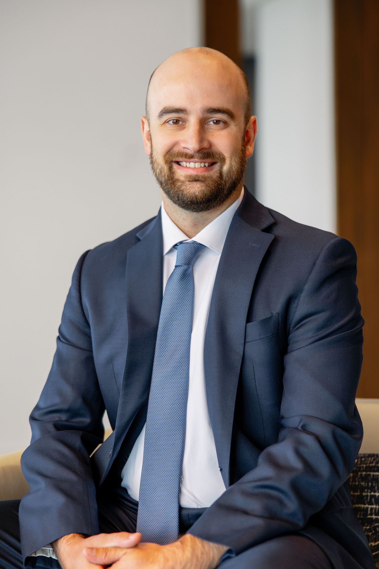 Professional bald man with beard in navy suit and tie sitting smiling in bright interior.