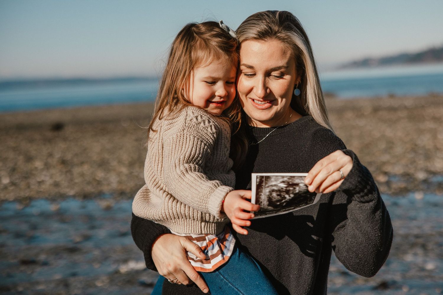 Mother holding toddler daughter while showing ultrasound photo at beach, announcing second pregnancy.