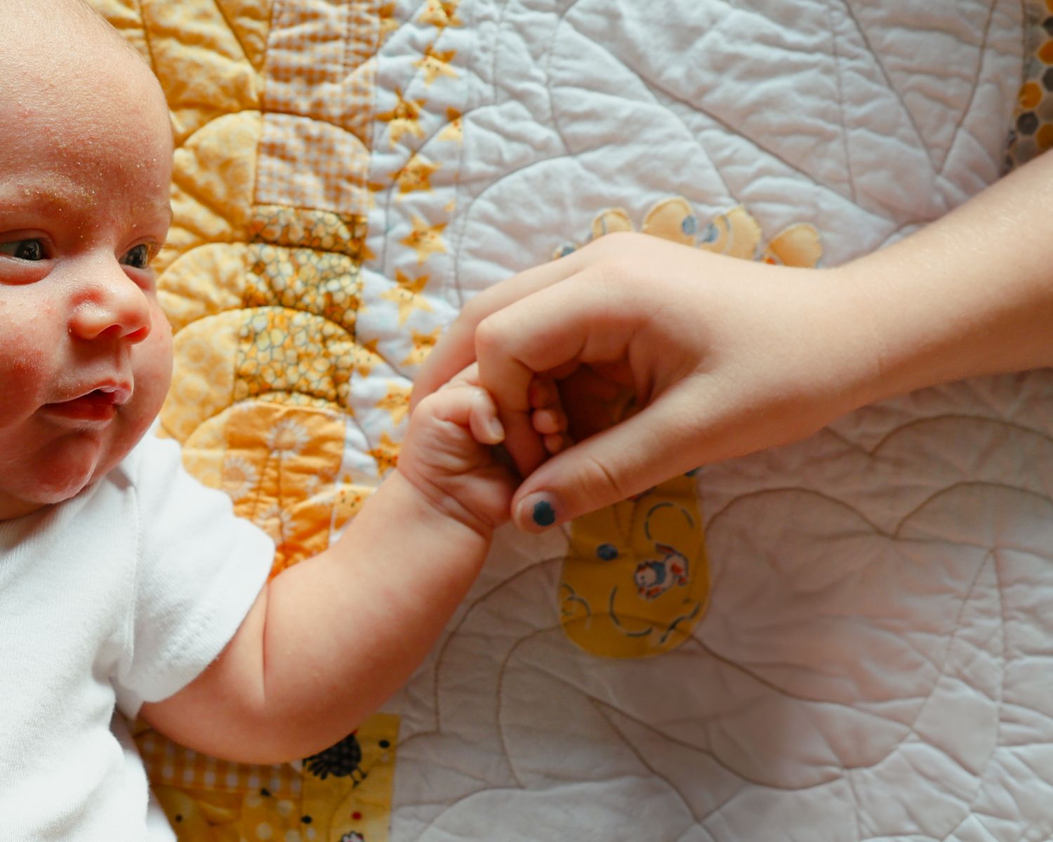 Close-up of newborn baby gripping an adult finger while lying on yellow quilted blanket.