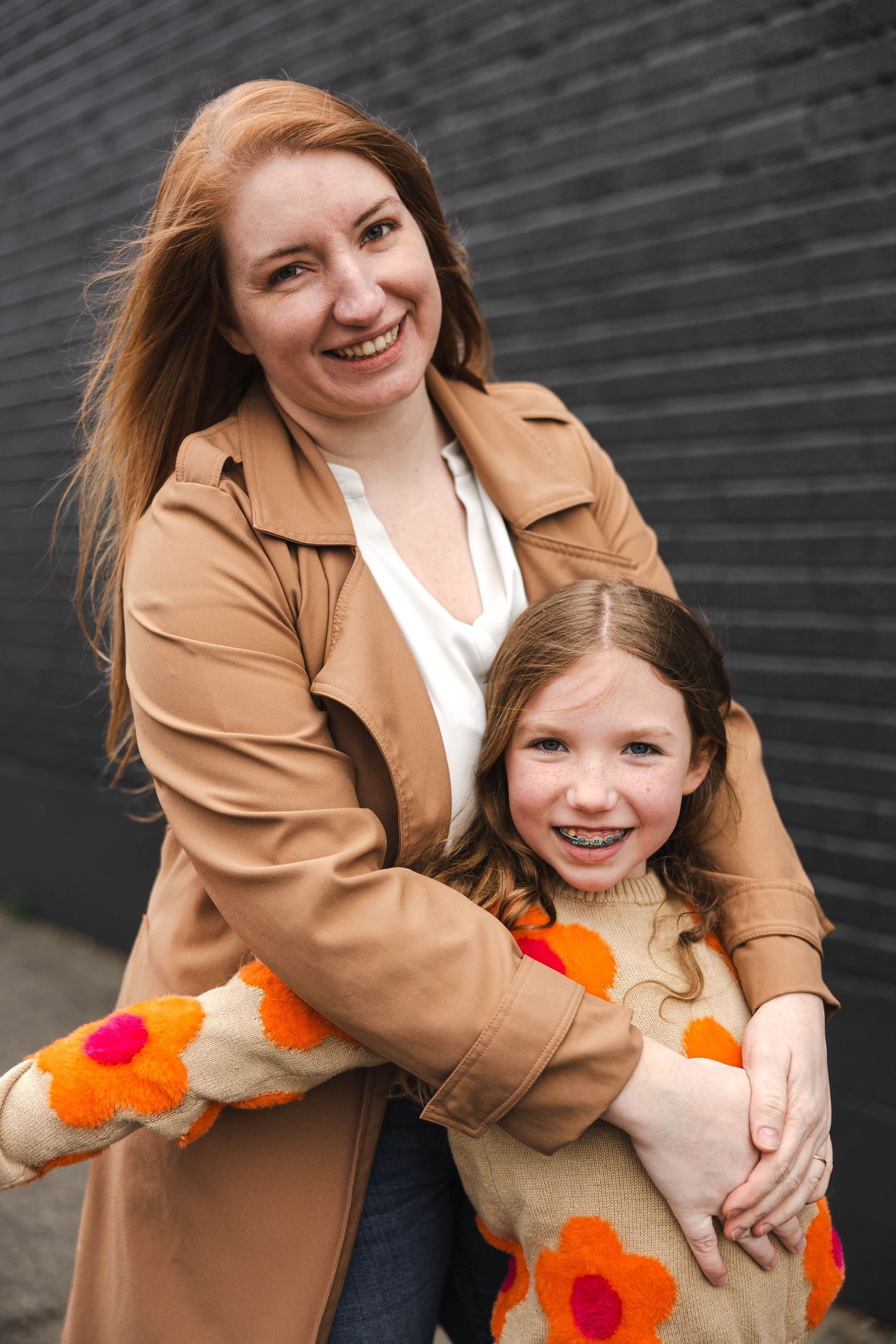 Smiling redhead mother hugging daughter in orange floral sweater against dark brick wall.