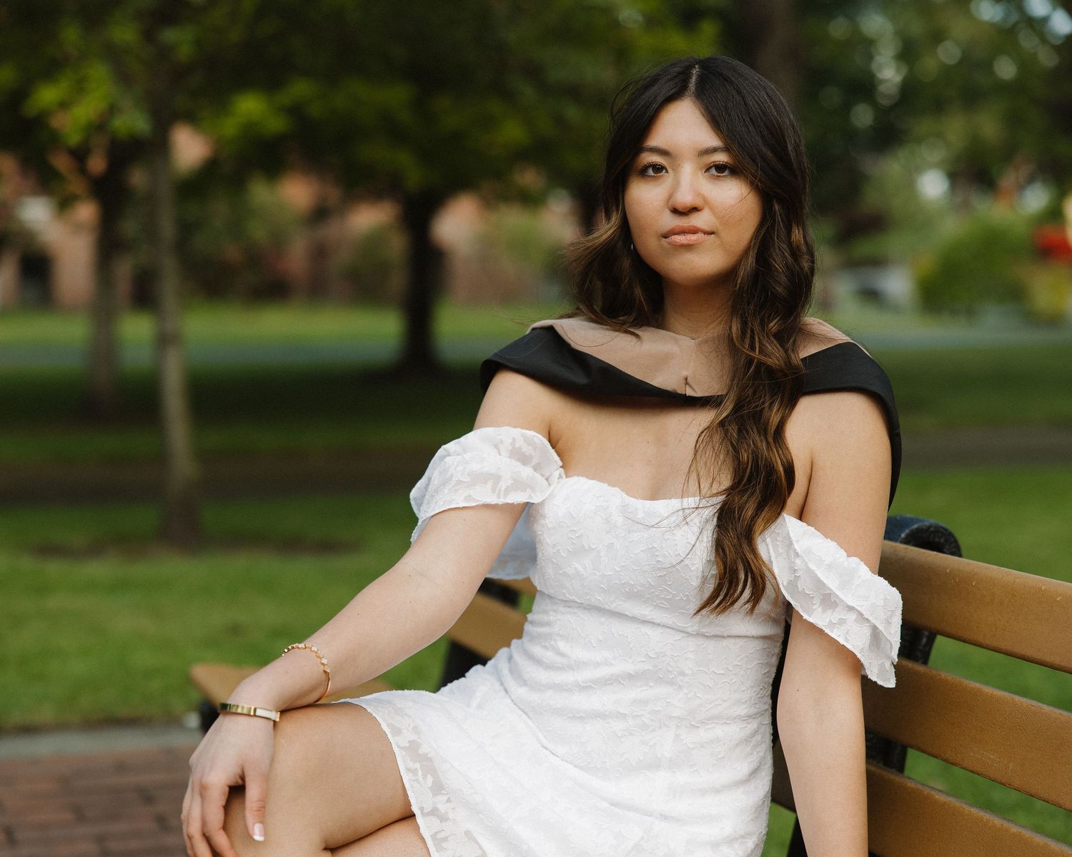 Young woman in white off-shoulder dress sitting on park bench outdoors.