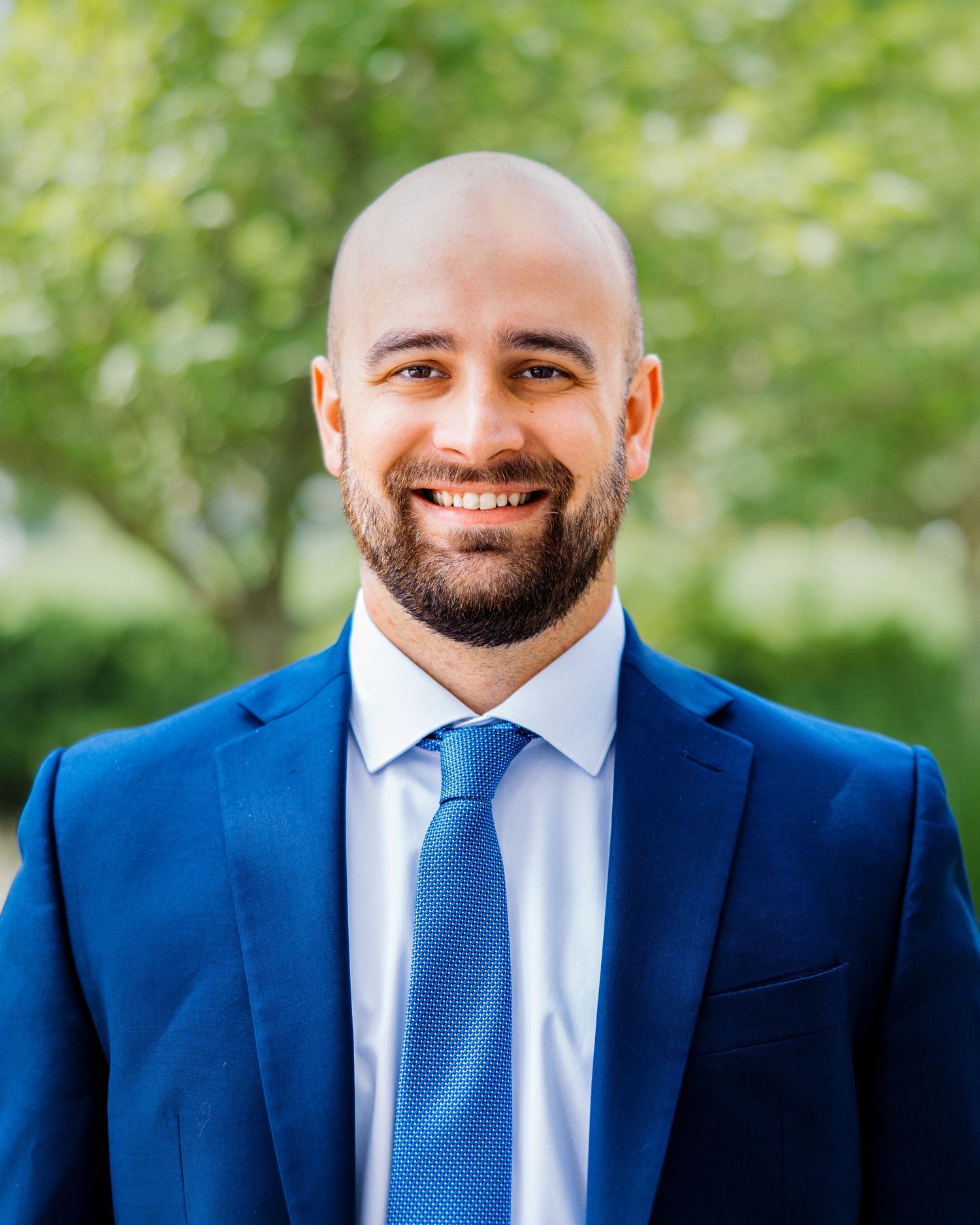 Smiling bald man with beard wearing blue suit and tie posing outdoors among green trees.