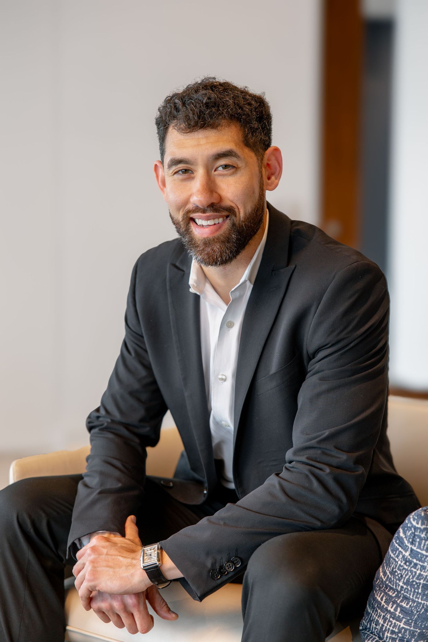 Smiling bearded man in dark suit seated casually in a modern office environment.