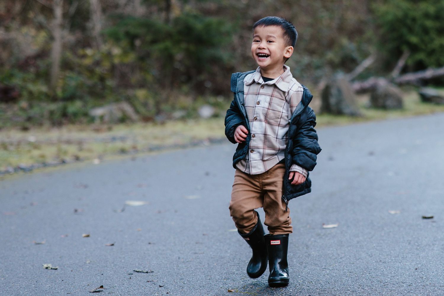 Happy young Asian boy running on a paved path outdoors, wearing a jacket, brown pants, and black boots.