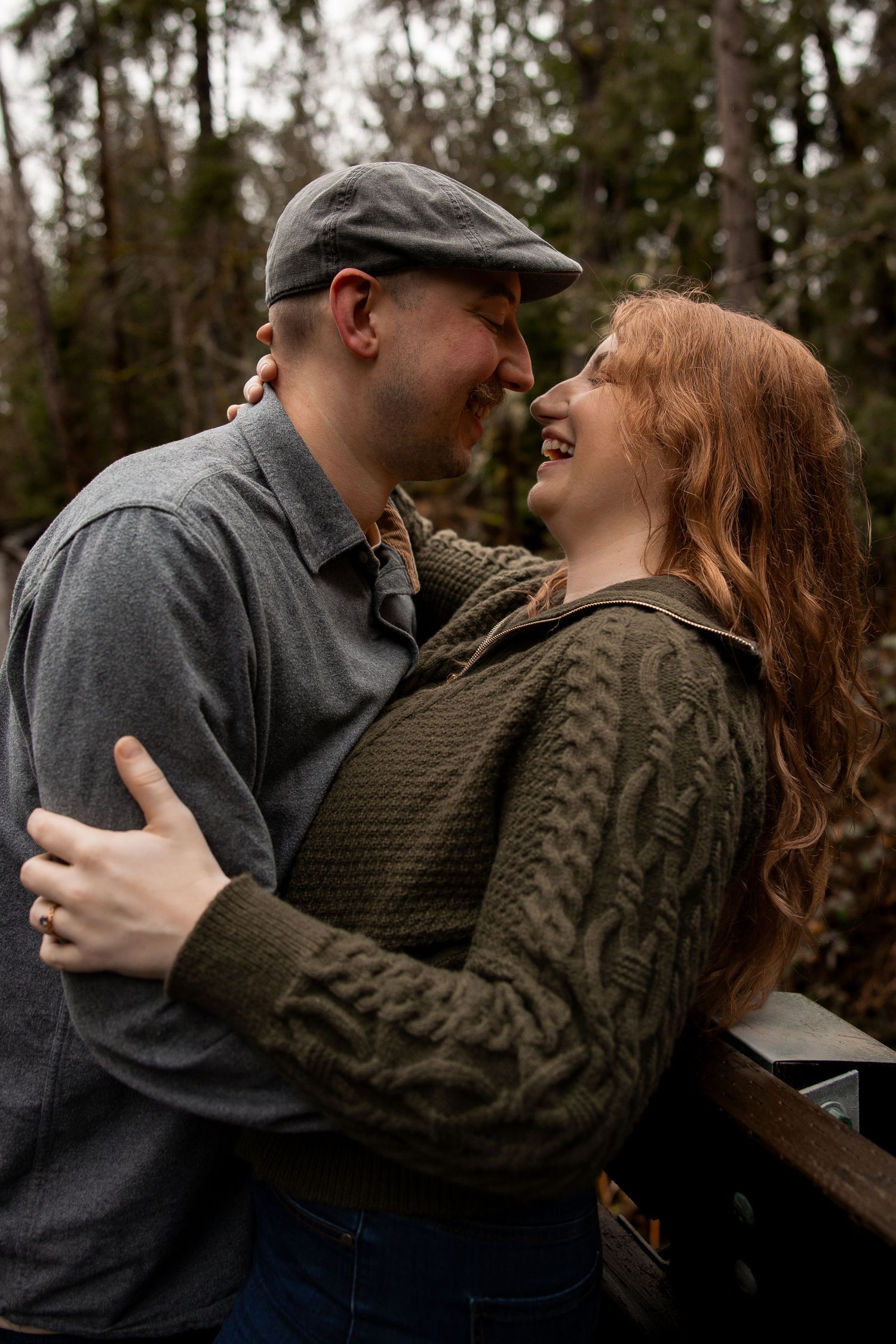 Smiling couple embracing closely outdoors, woman with red hair in green sweater, man in gray cap.