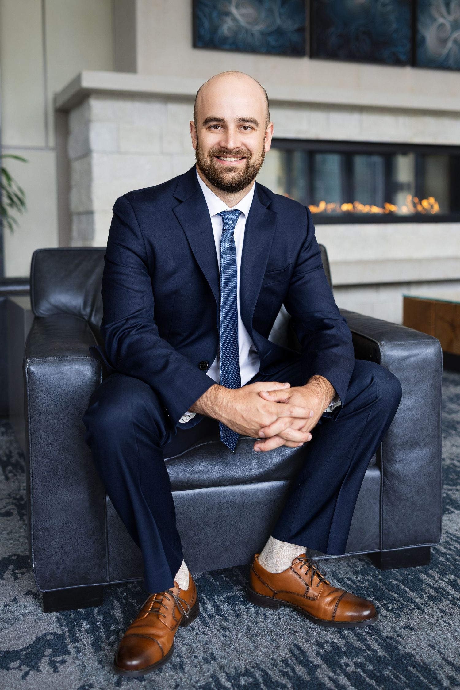Bald bearded man in navy suit and blue tie seated smiling in modern lobby with fireplace background.