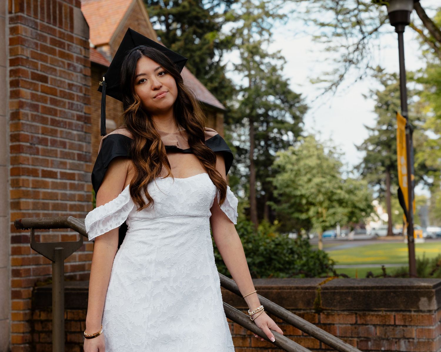 Female graduate in white dress and black graduation cap poses gracefully on brick steps beside a brick building.