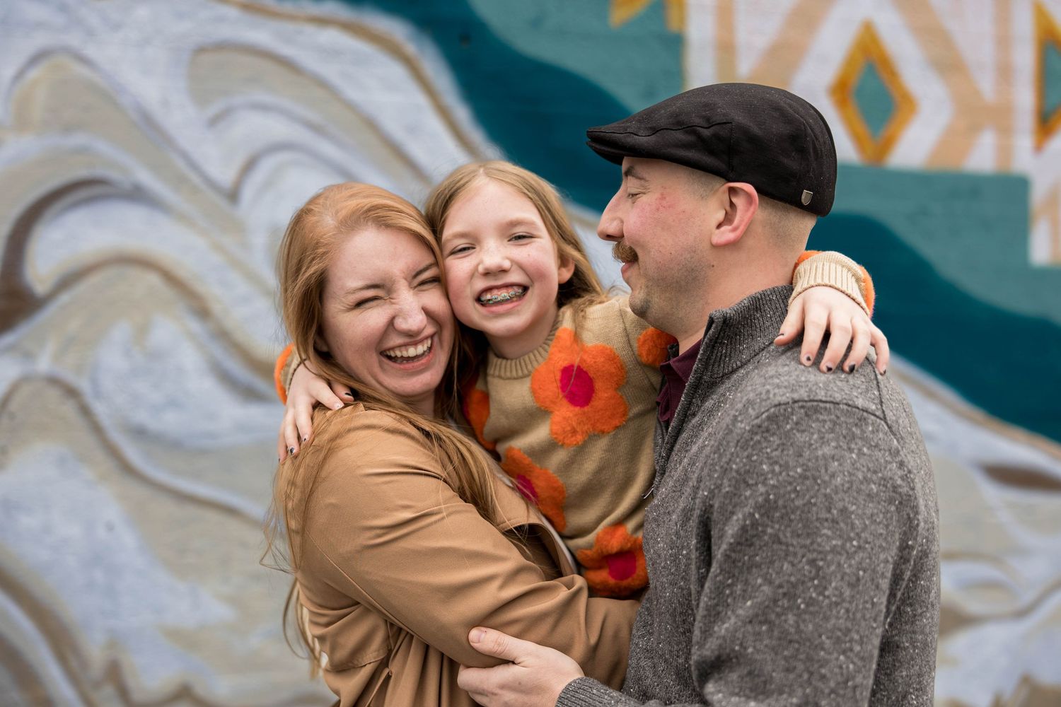 Happy family of three hugging and laughing in front of colorful mural outdoors.
