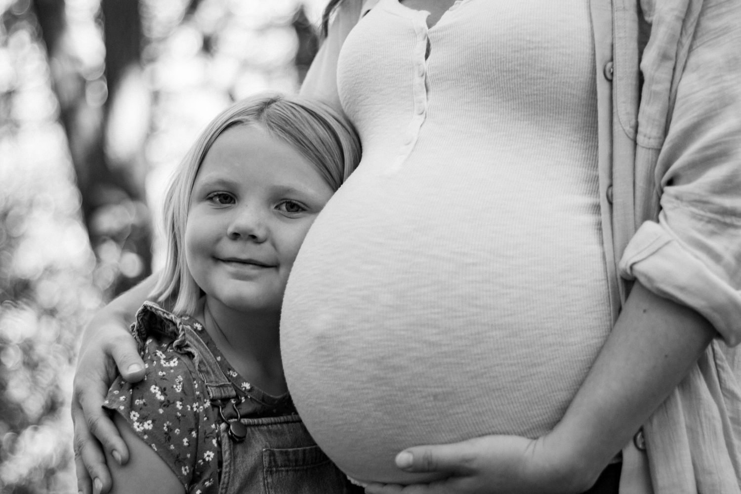 Black and white photo of a young girl hugging a pregnant womans belly outdoors.
