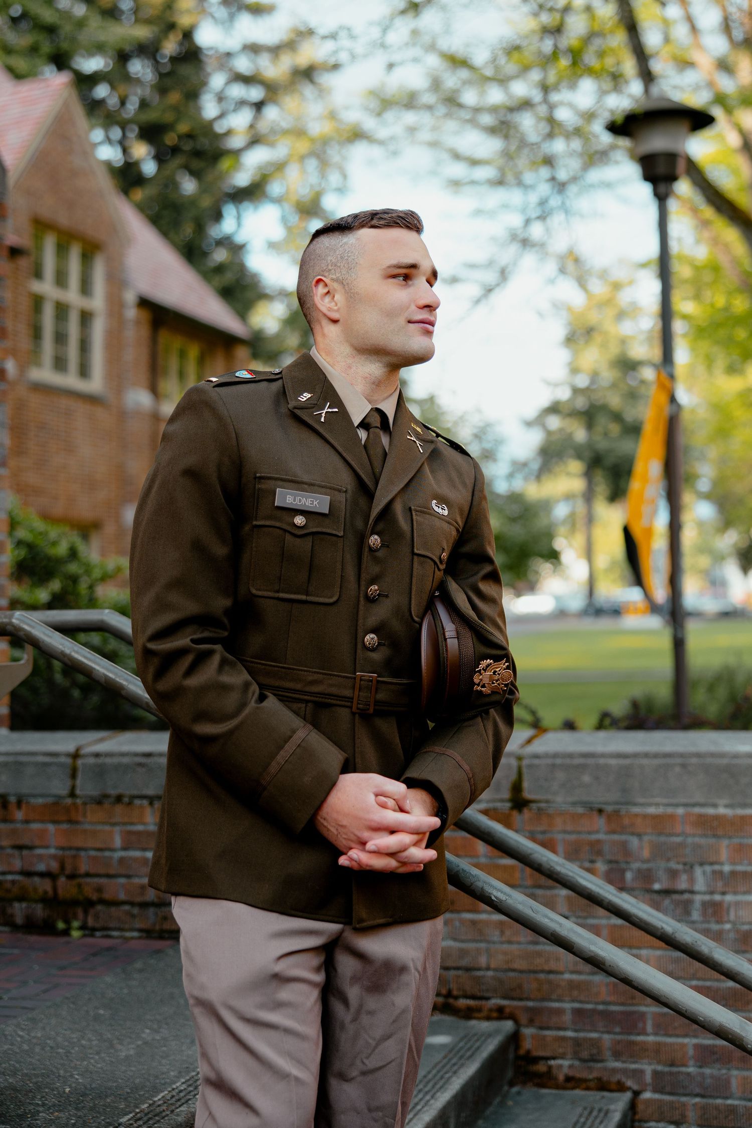 Young military officer in olive dress uniform stands confidently outdoors near brick building and green trees.