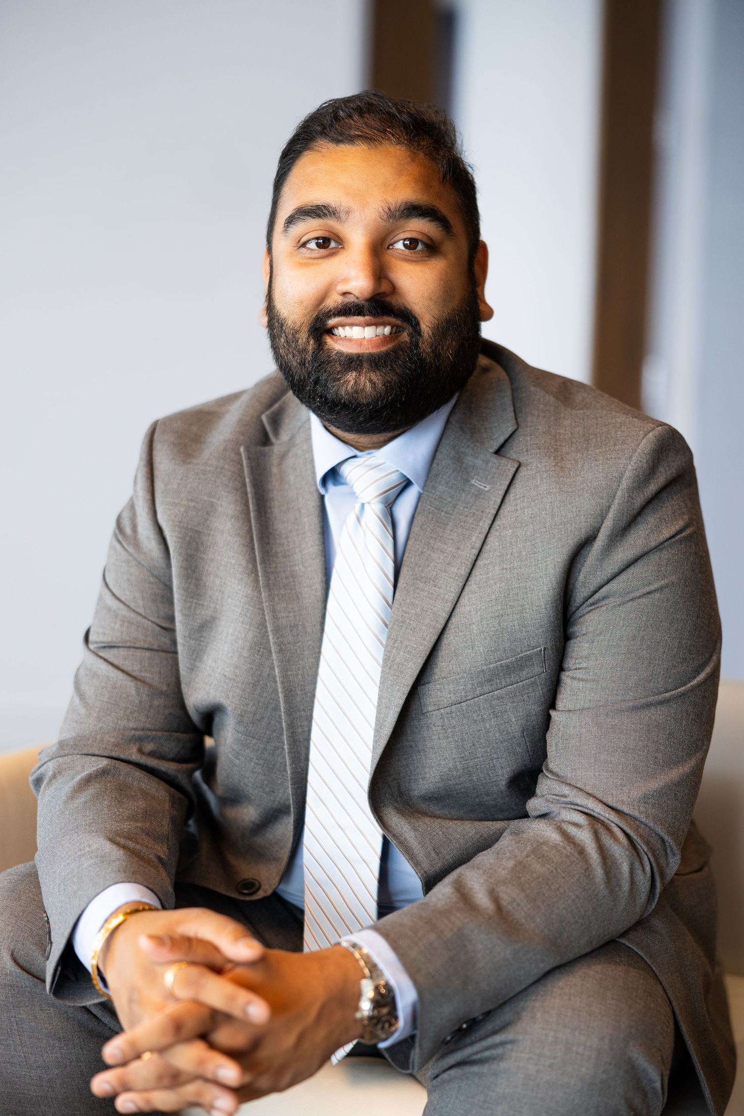 Smiling bearded man in gray suit and striped tie seated casually in bright modern office environment.